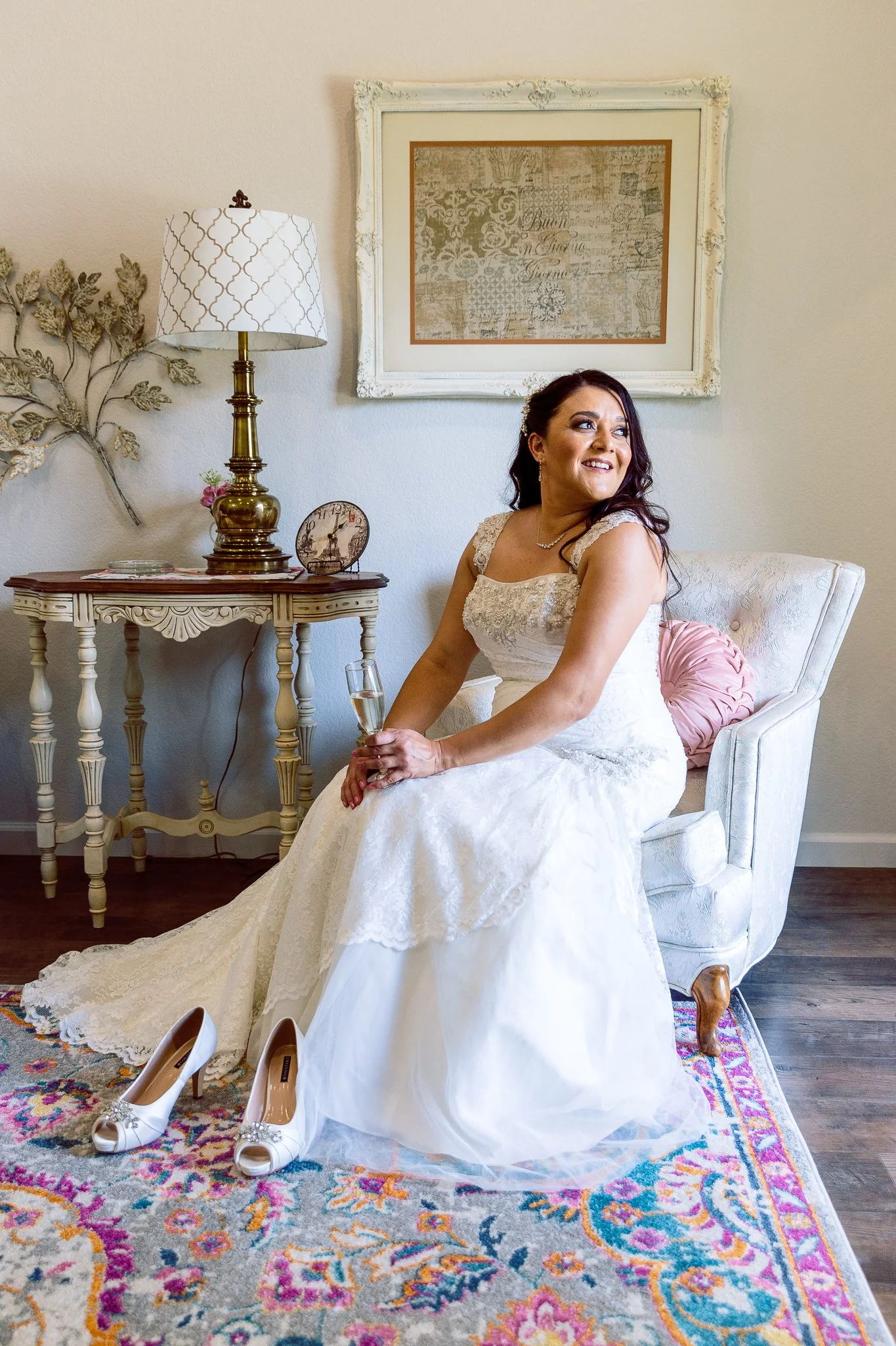 A bride sitting on a white armchair in a cozy room, holding a glass of champagne, wearing a white wedding dress, with her white high heels placed on a colorful rug in front of her. There is a side table with a lamp, a clock, and decorative items behi