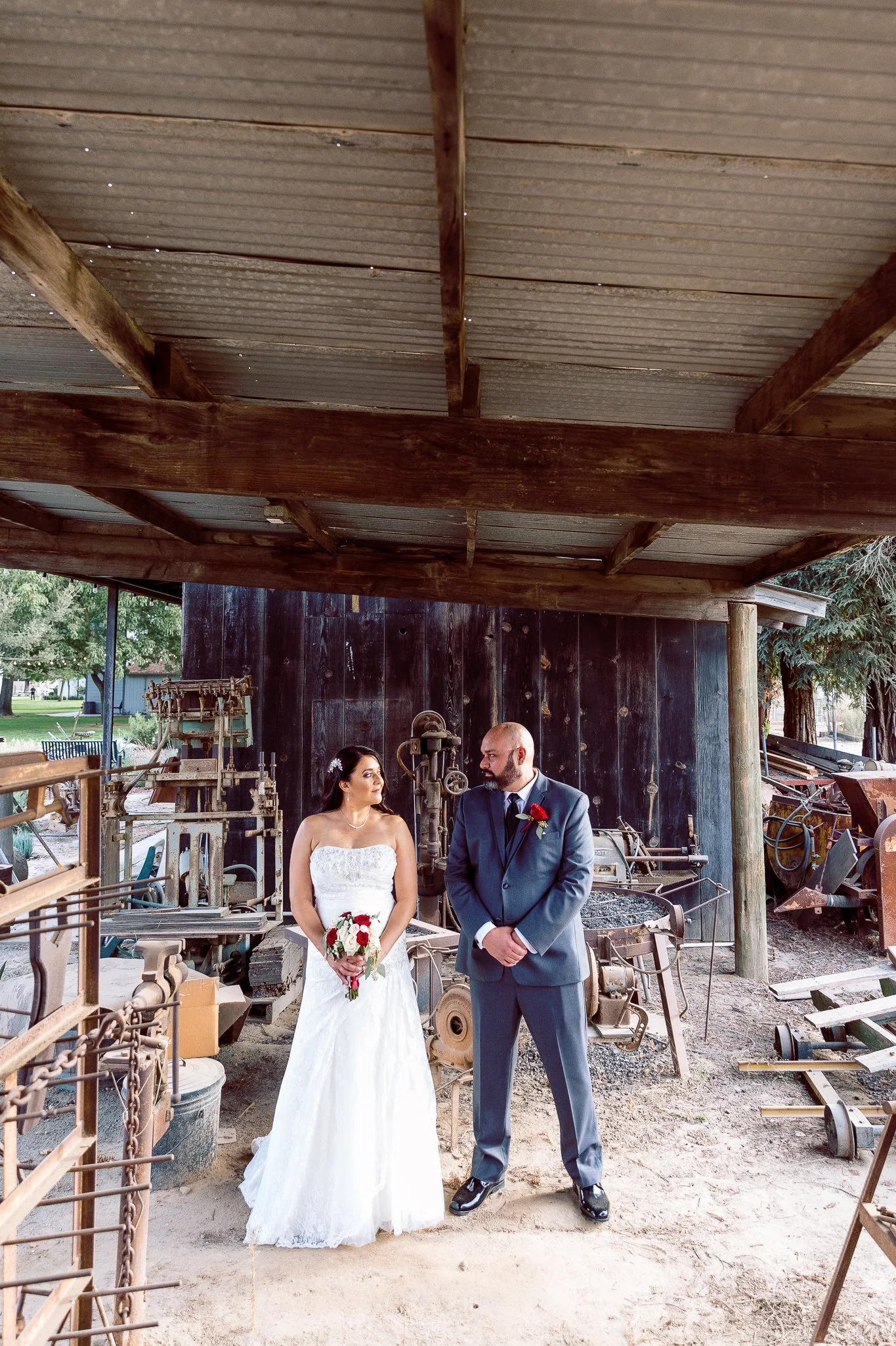 A bride and groom standing under a rustic outdoor shelter, facing each other, with vintage machinery and tools in the background.