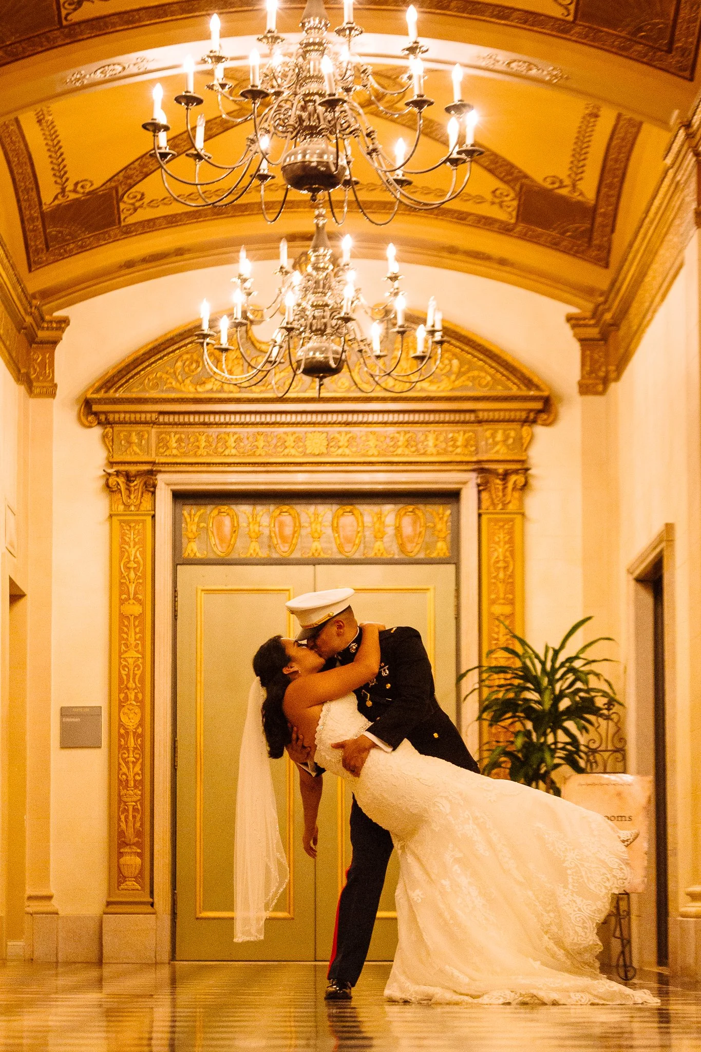 A couple dressed in wedding attire sharing a kiss in an elegant grand hall with ornate gold detailing and chandeliers.