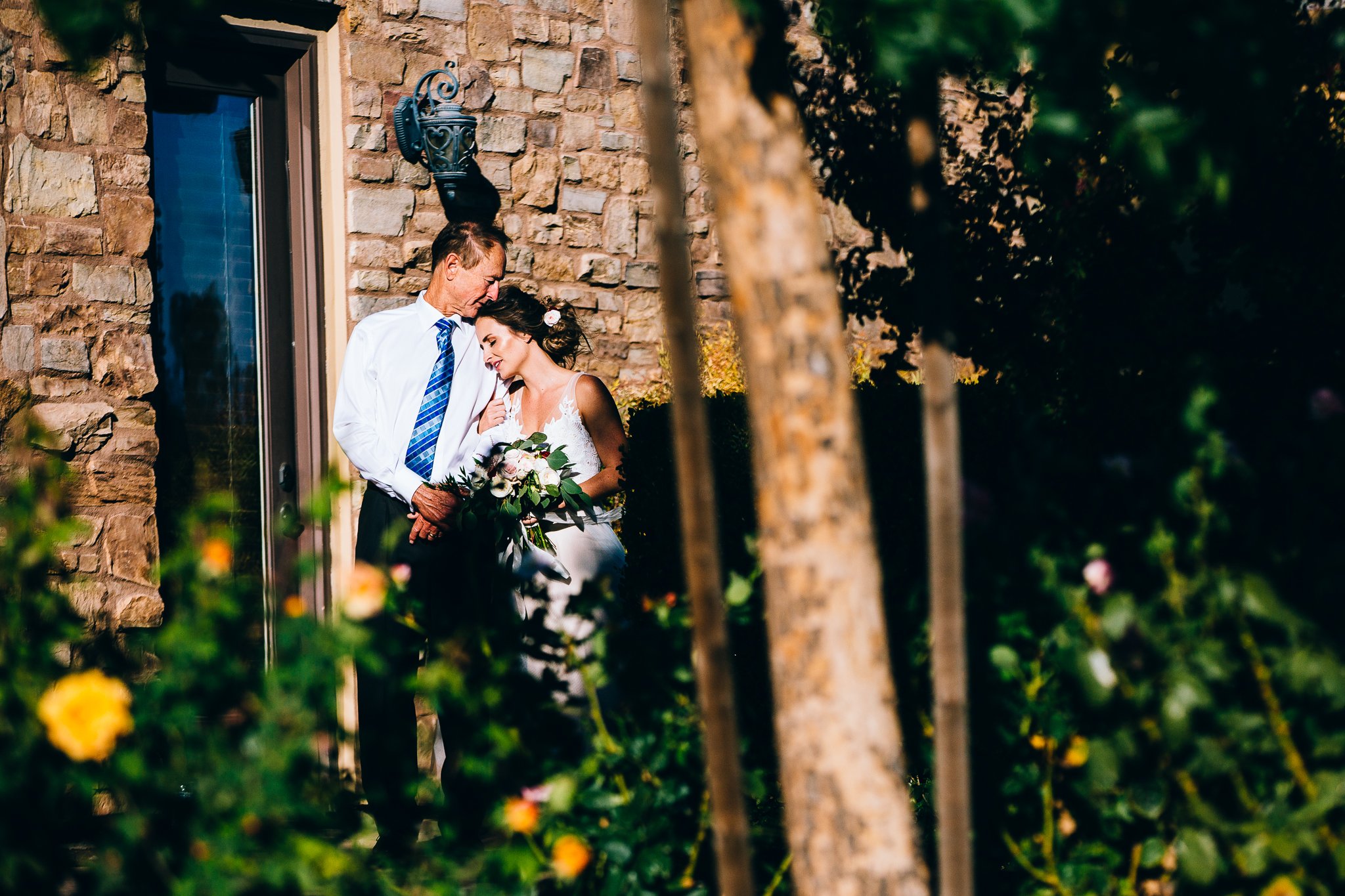 An elderly man and a young woman in a wedding dress standing close together outside beside a brick wall, with greenery and flowers around. The woman is holding a bouquet of flowers and the man is affectionately leaning towards her, both appear to be 