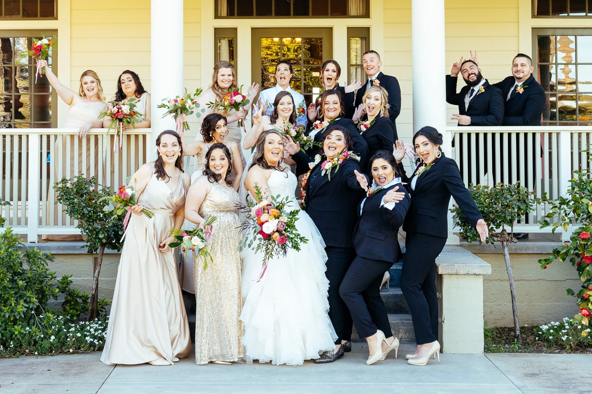 Wedding party group photo on front steps of a house, including bride, groom, bridesmaids, and groomsmen celebrating and posing cheerfully.