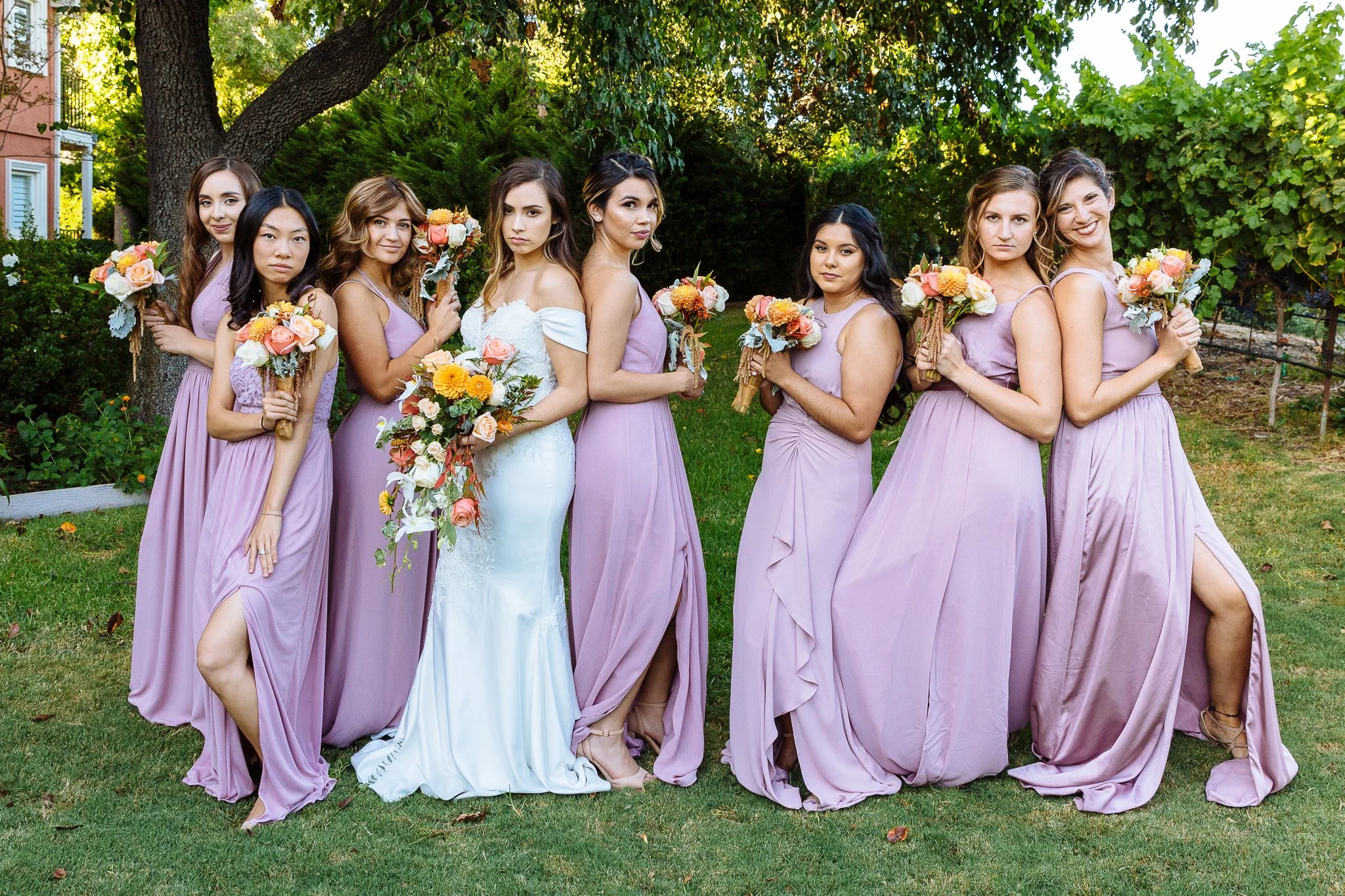 A bridal party of nine women standing outdoors in a garden with green trees and grass, dressed in lavender bridesmaid dresses, except for the bride in a white wedding gown, holding bouquets of flowers.
