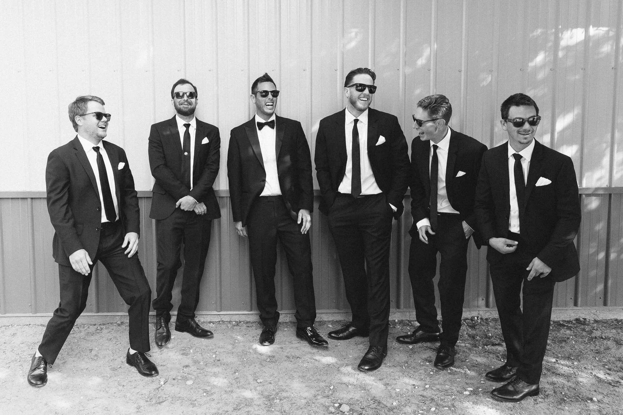 Group of seven men in black tuxedos and sunglasses standing outdoors against a metal building, smiling and laughing.