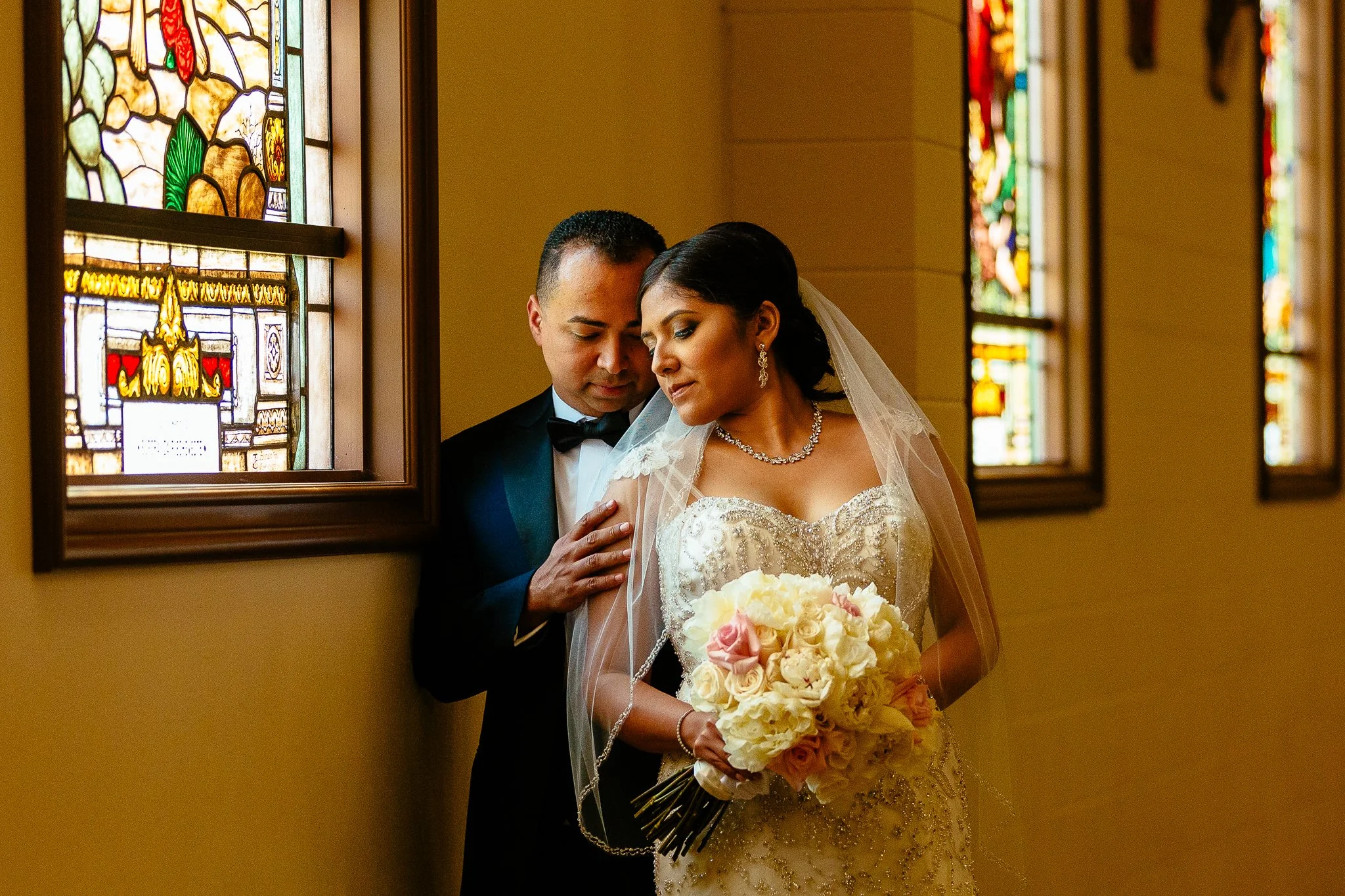 A bride and groom standing close together inside a church, with stained glass windows in the background. The bride is holding a bouquet of cream and pink roses, wearing a detailed wedding dress and veil. The groom is dressed in a tuxedo with a bow ti