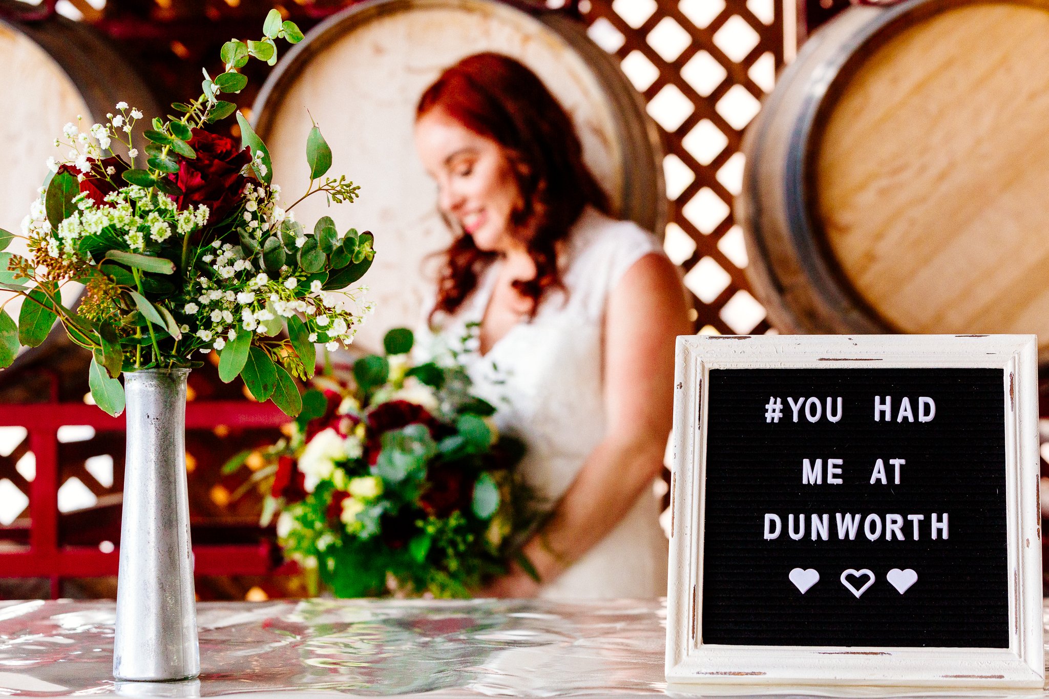 A woman with brown hair wearing a white dress, holding a bouquet of flowers, standing behind a table with a vase of flowers and a sign that reads 'You had me at Dunworth' with hearts. The background features large wooden barrels and lattice woodwork.