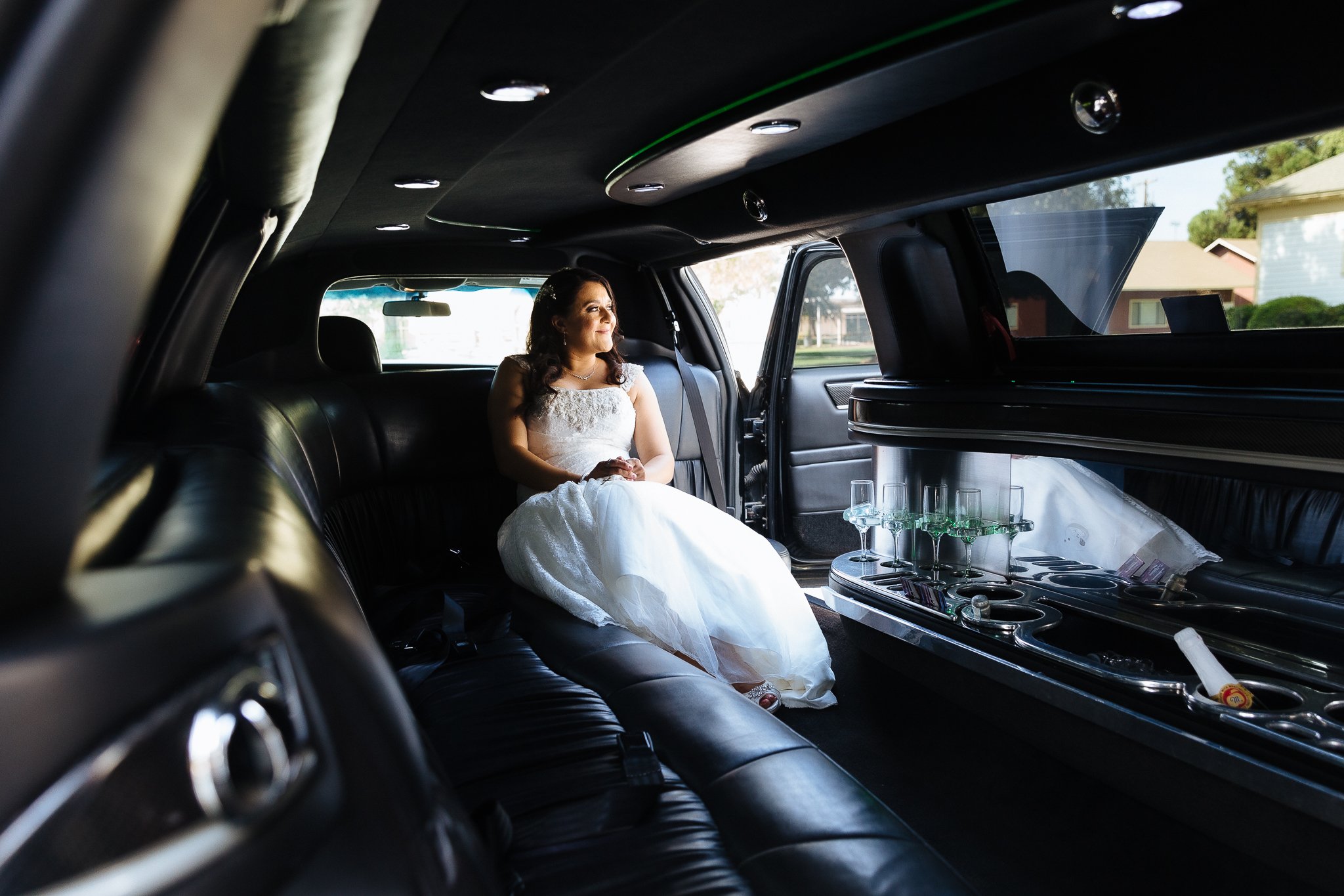 A woman in a white wedding dress sitting inside a black limousine, looking out the window with a smile.