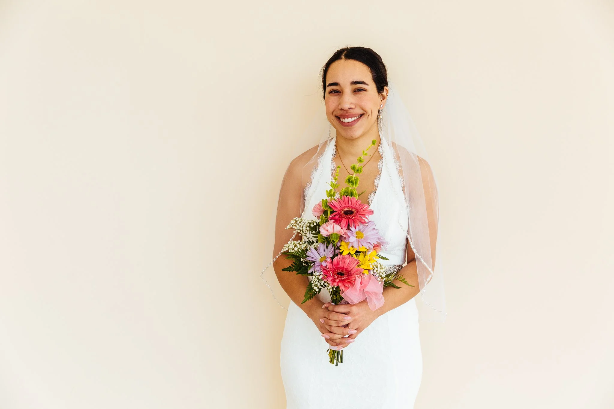 Young woman in a white wedding dress holding a colorful bouquet of flowers, smiling, against a plain beige background.