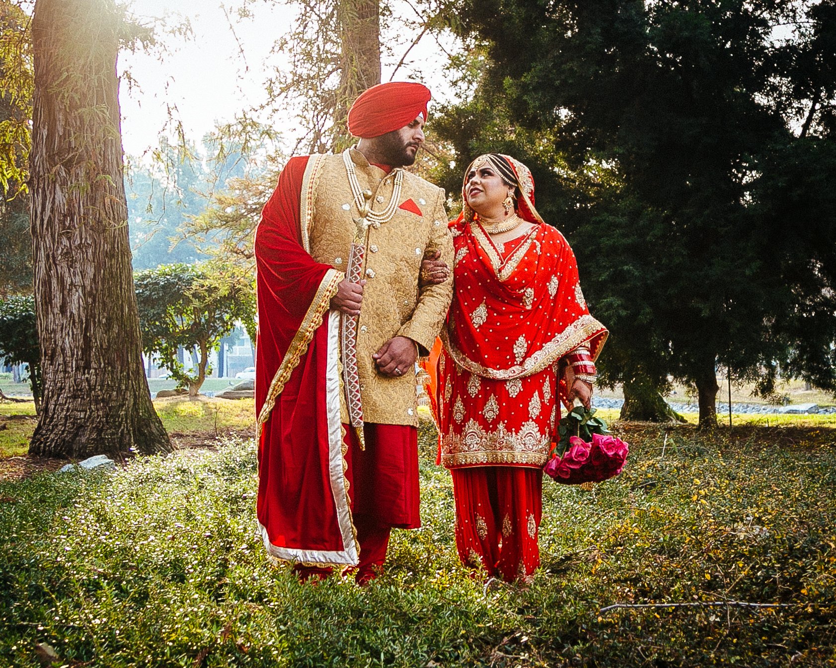 A couple dressed in traditional Indian wedding attire standing in a green park. The groom is wearing a gold sherwani with red accents and a red turban. The bride is wearing a red and gold embroidered saree, jewelry, and holding a bouquet of pink rose