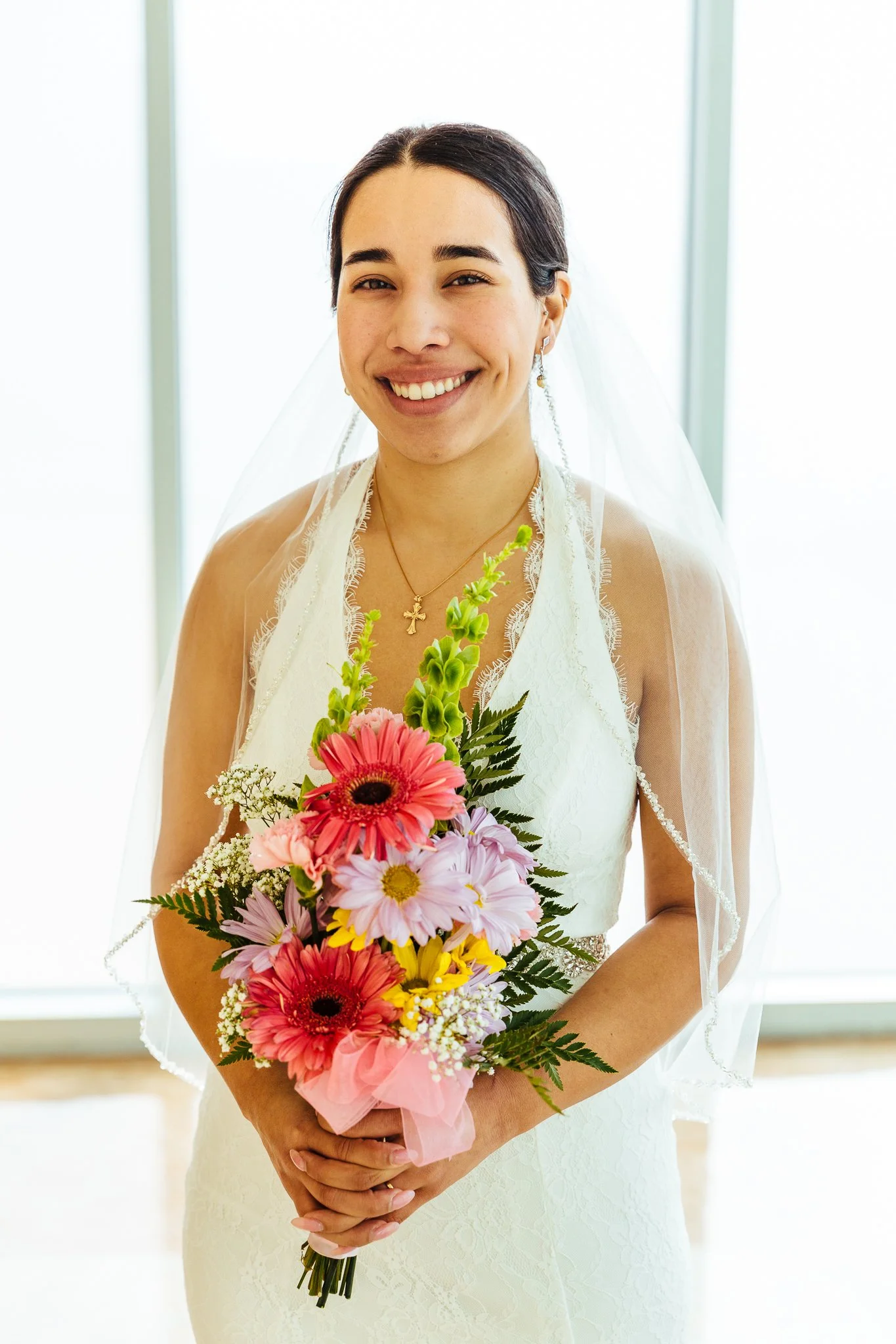 A bride in a white wedding dress and veil holding a bouquet of flowers, smiling indoors with bright background.
