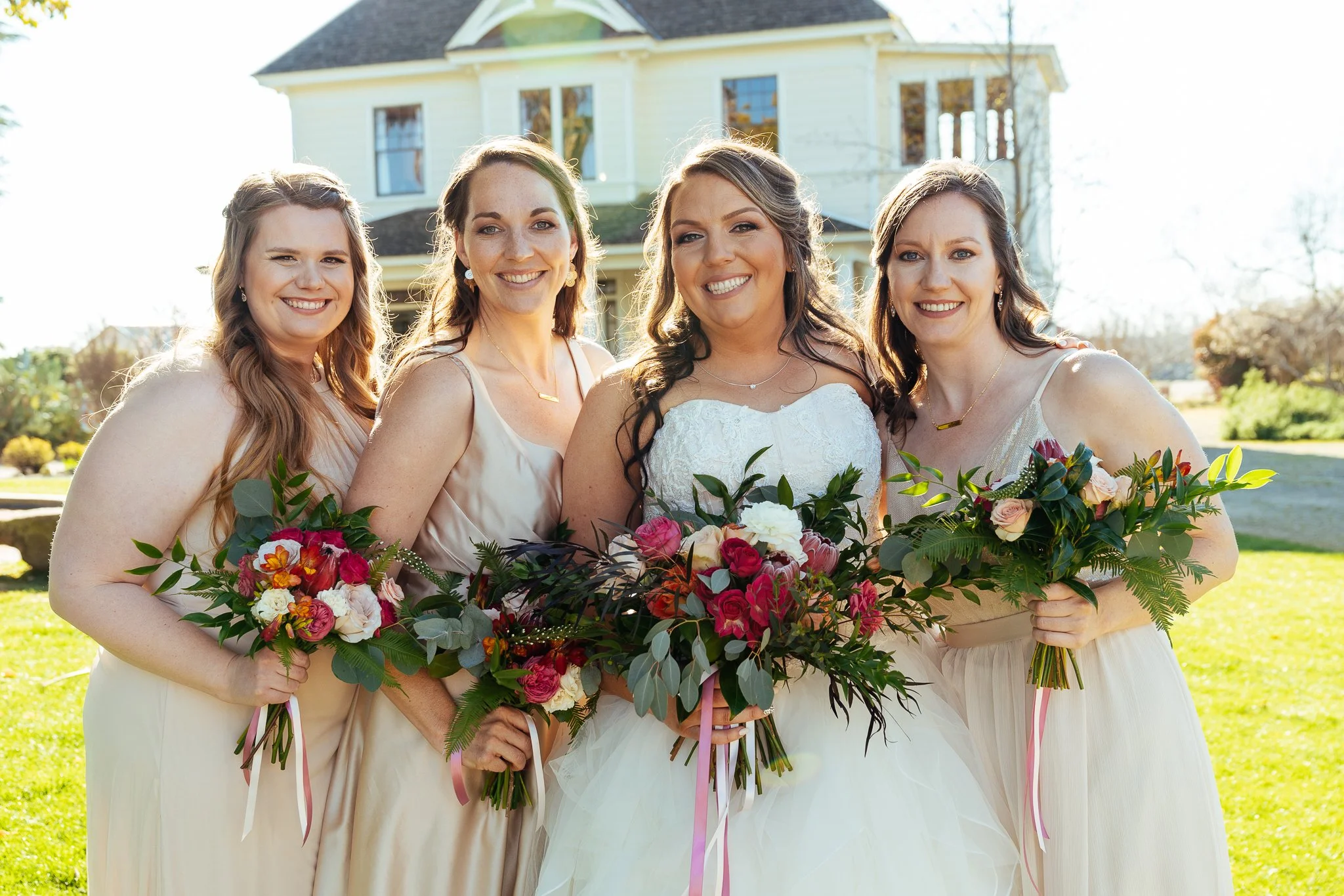 Bride and her bridesmaids standing outdoors in front of a house, holding bouquets of pink, white, and red flowers with greenery, smiling at the camera.