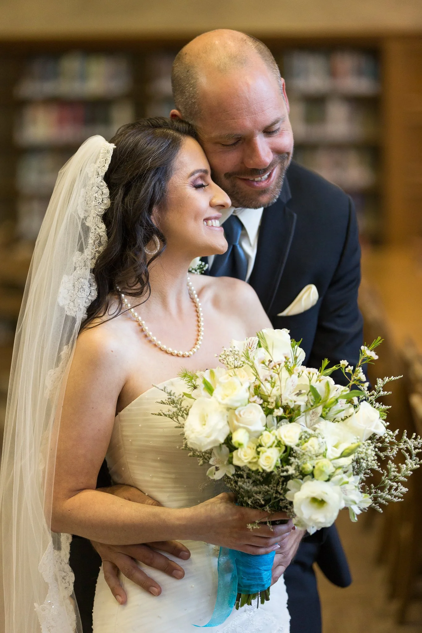 A bride and groom smiling and hugging at their wedding, holding a bouquet of white flowers, with a background of bookshelves.