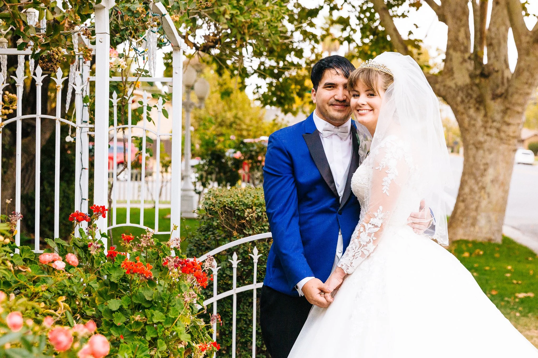 A bride and groom in wedding attire standing outdoors, holding hands and smiling, with a white garden fence, trees, and flowers in the background.