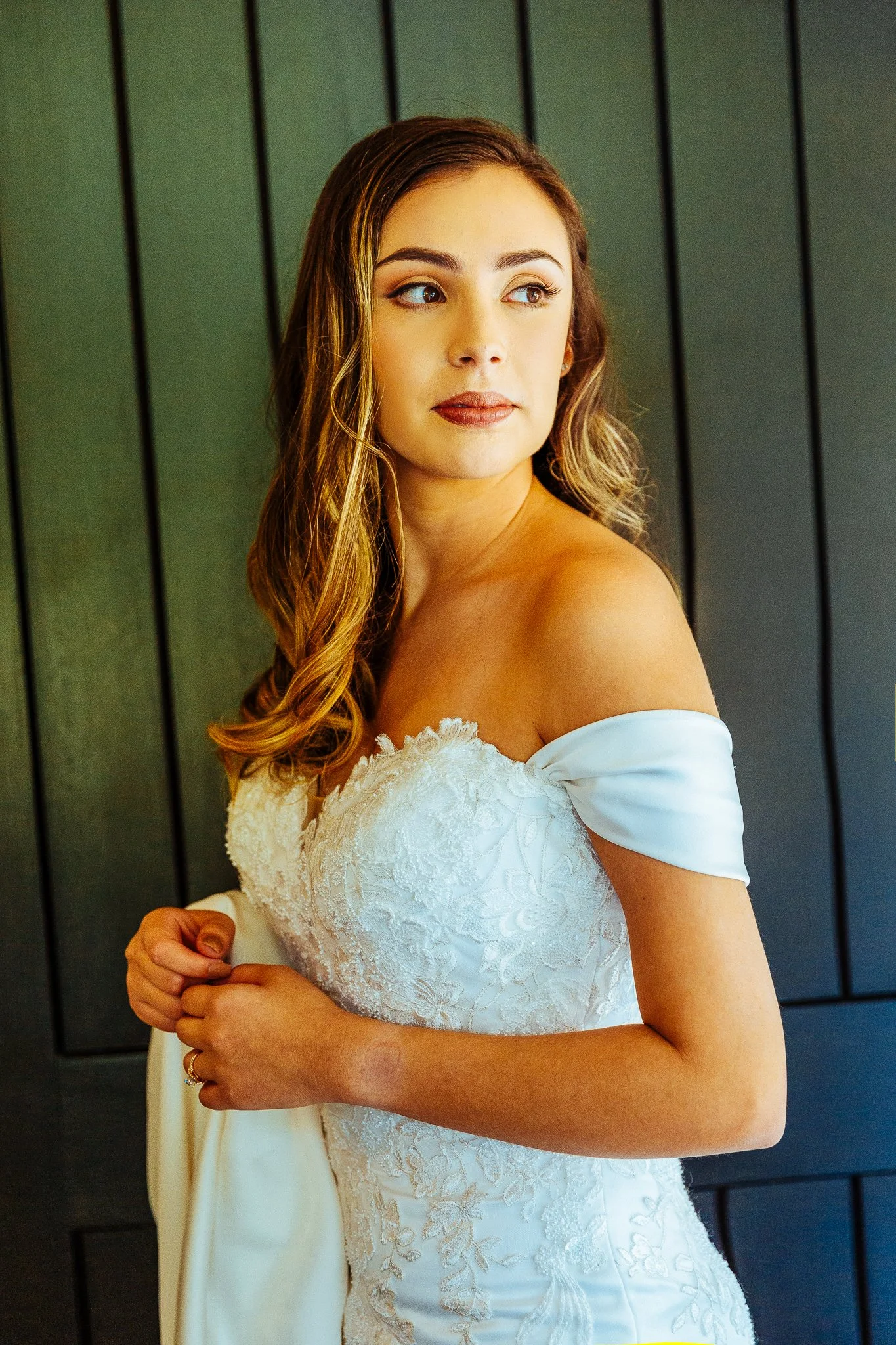 A young woman in an off-shoulder white wedding dress with lace details, looking to the side with a thoughtful expression, standing against a black paneled wall.