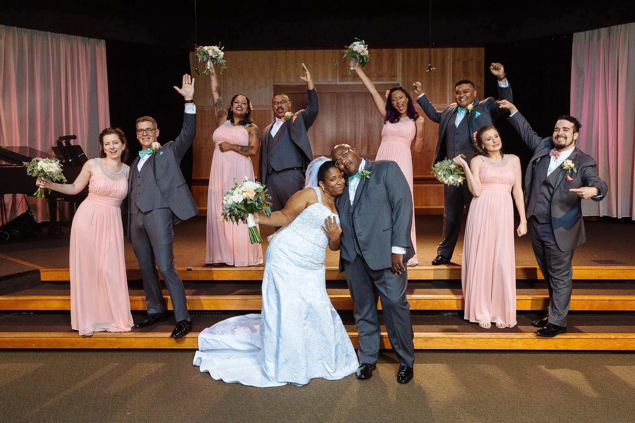 A group of wedding party members, including bride and groom, celebrating on a stage with pink bridesmaids' dresses and grey suits, holding bouquets, with a piano and curtains in the background.