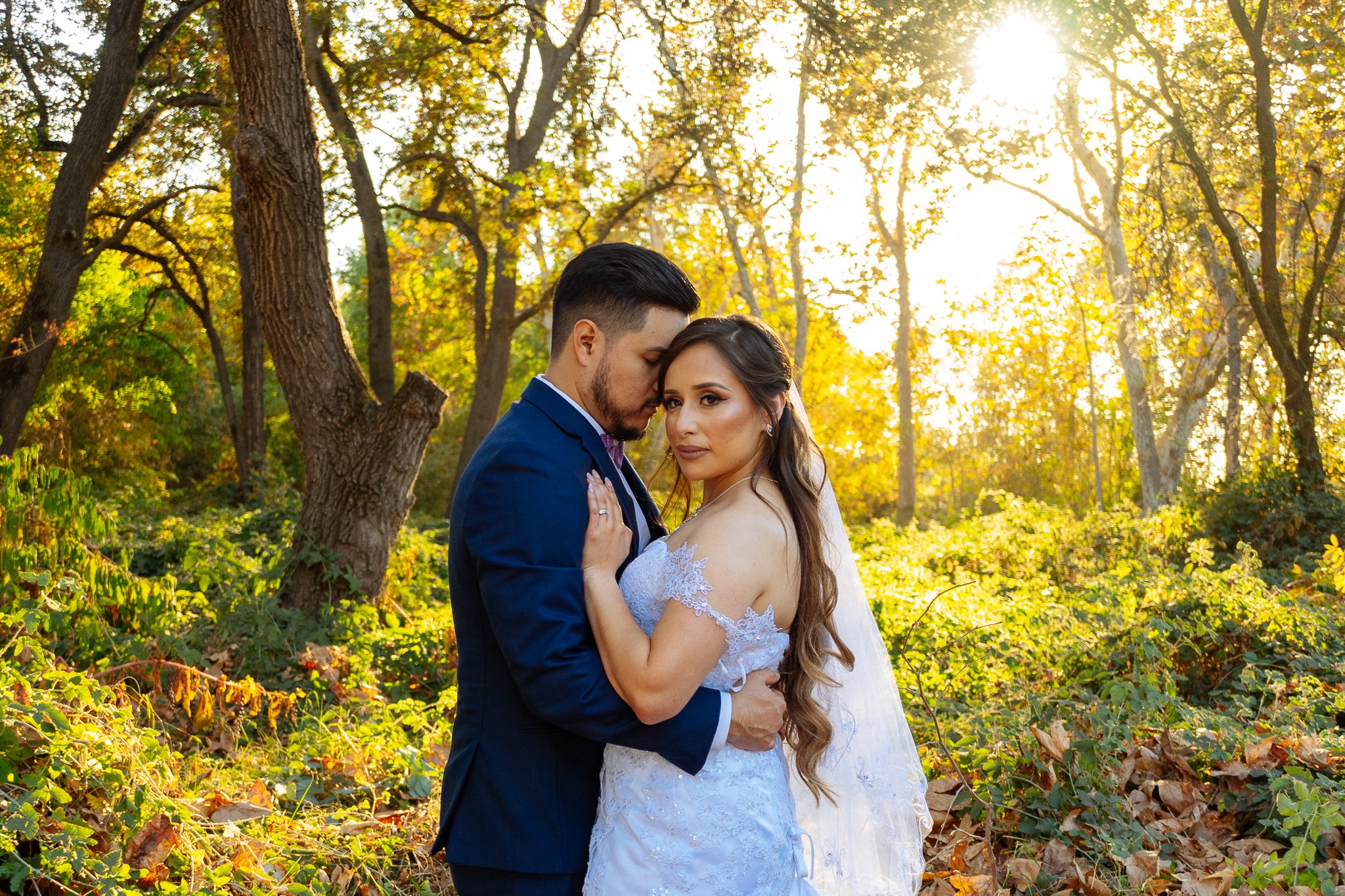 A bride and groom embrace in a forest during sunset, with the bride in a white wedding dress and the groom in a navy blue suit, surrounded by autumn foliage.