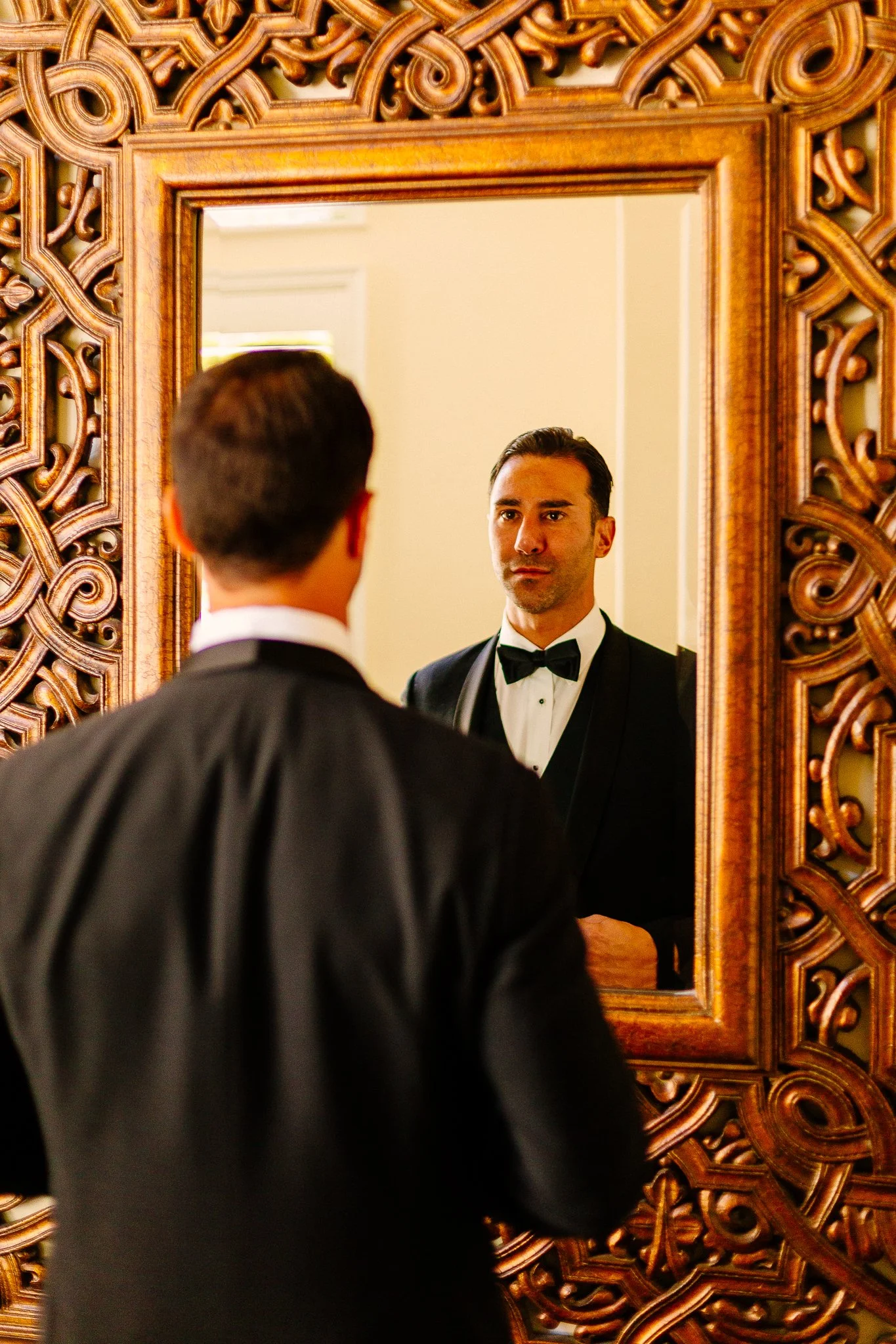 A man in a tuxedo looking at his reflection in a decorative wooden-framed mirror.
