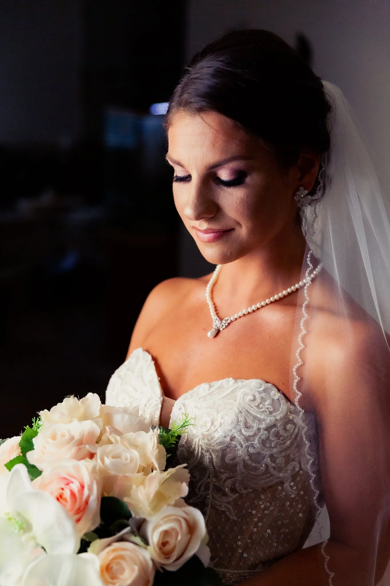 A bride with dark hair styled up, wearing a white wedding dress with lace detailing, a pearl necklace, and earrings, holding a bouquet of light pink and white roses, with a soft smile and looking down.