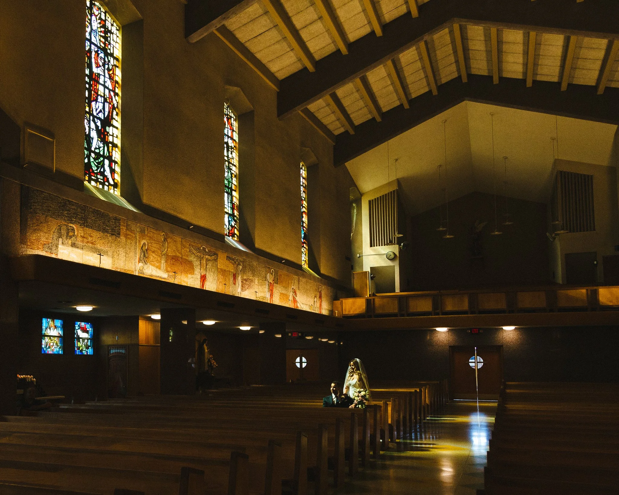 Interior of a church with wooden pews, stained glass windows, and a high ceiling. A bride and groom sit in the front pew, with other guests visible in the background.