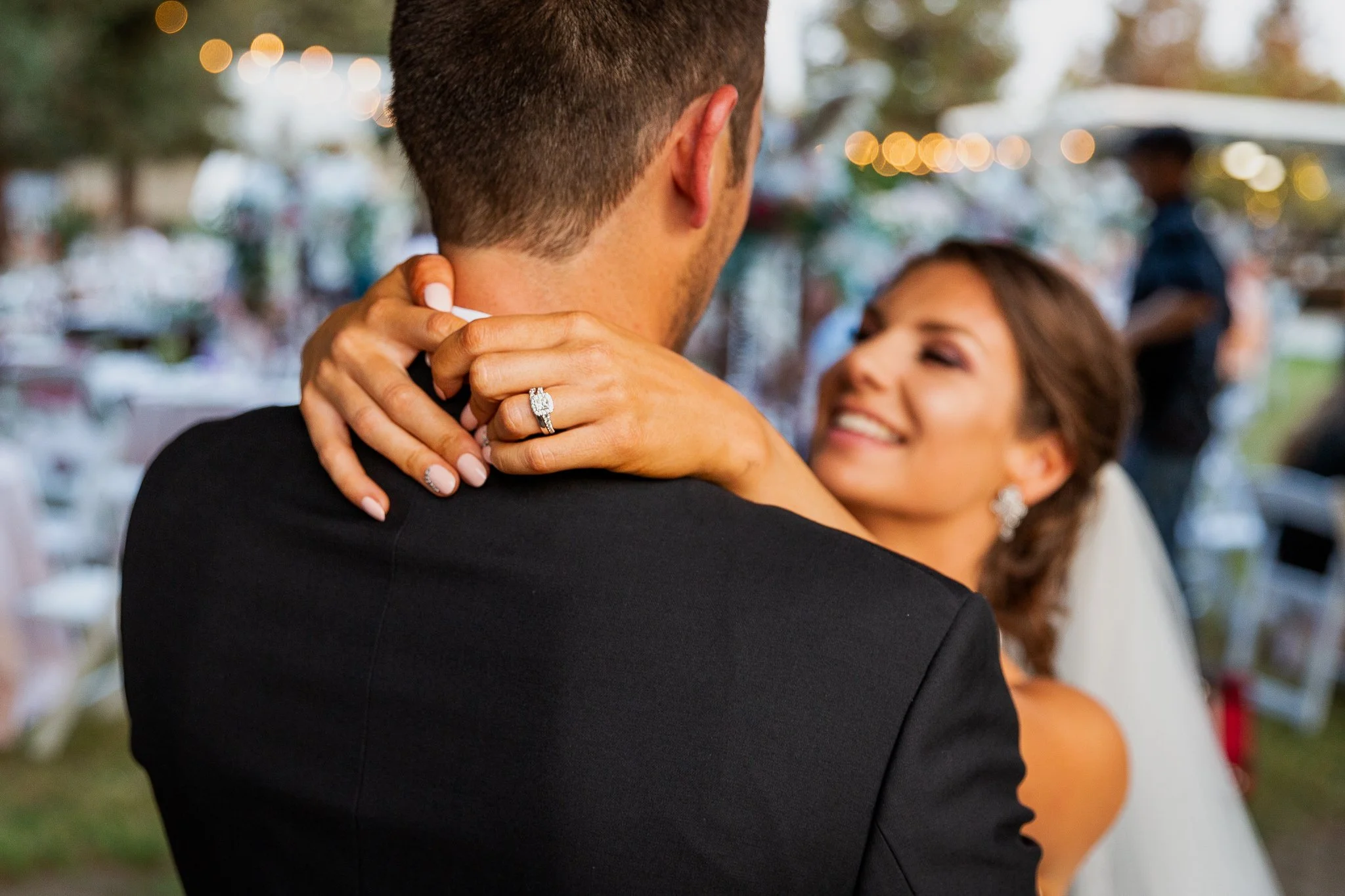 A bride and groom sharing a hug at their wedding reception, with the bride smiling and showing her wedding ring.