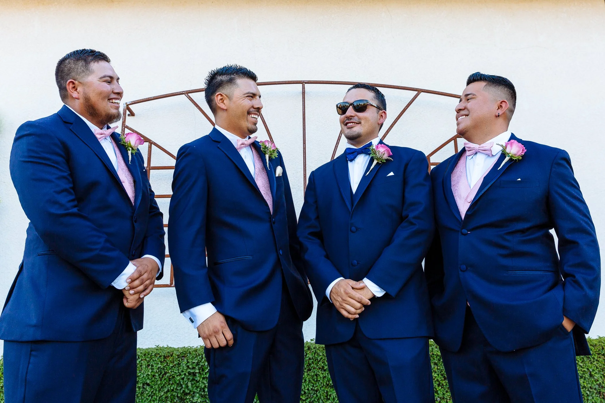 Five men dressed in navy suits and pink or blue bow ties standing outdoors, smiling and talking to each other, with a white wall and greenery in the background.