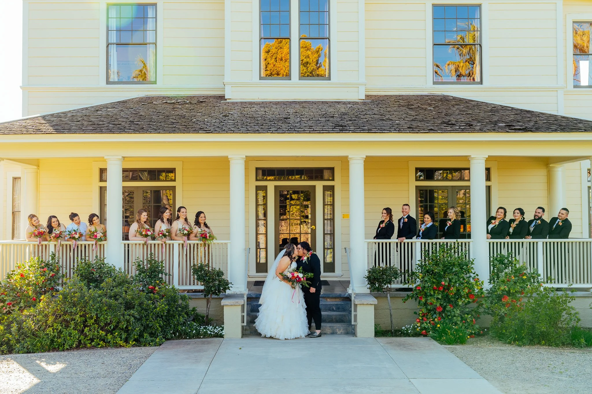 Wedding couple kissing on front steps of a yellow house with wedding party members lined up on porch, holding bouquets, during daytime