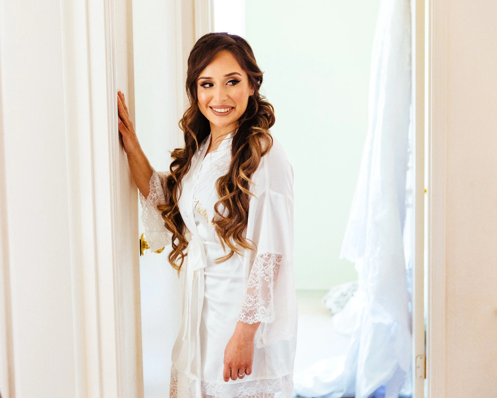 A woman with long, curly brown hair in a white robe with lace sleeves, smiling and standing in a doorway, preparing for her wedding day.