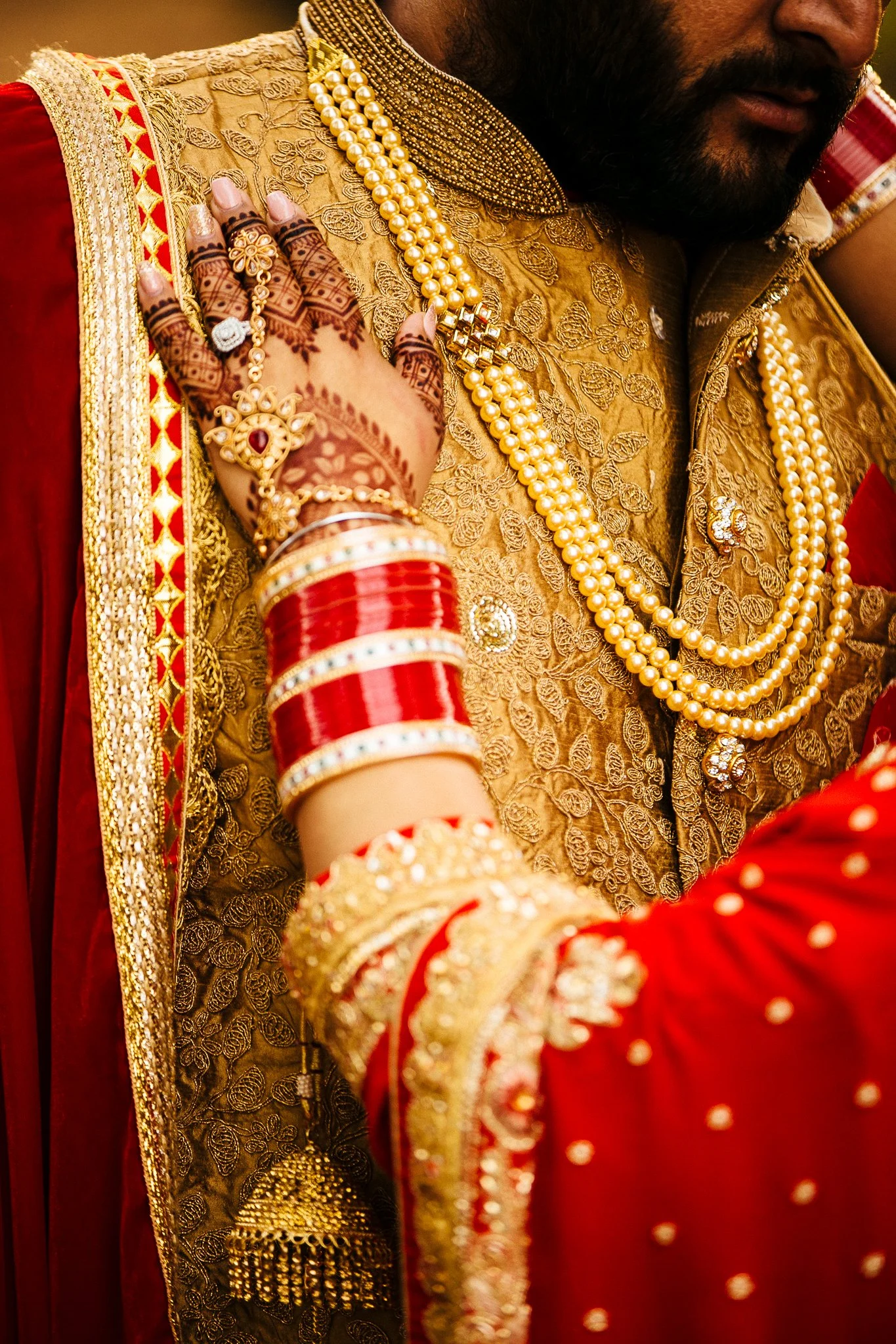 Close-up of a groom dressed in traditional Indian wedding attire with intricate gold embroidery, layered pearl necklaces, red and gold bangles, and ornate jewelry.