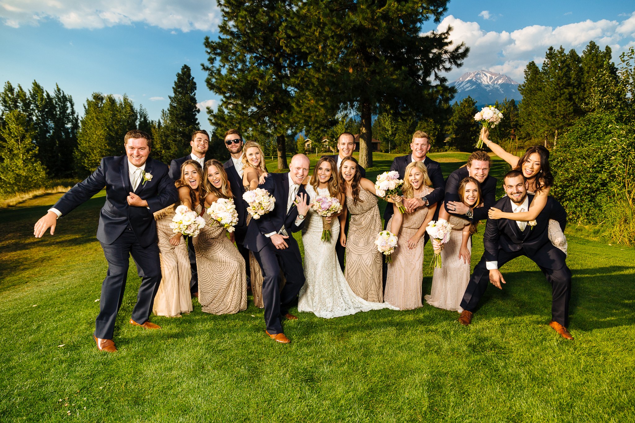 Group of wedding party members in formal attire, smiling and posing on a grassy field with trees, mountains, and a partly cloudy sky in the background.