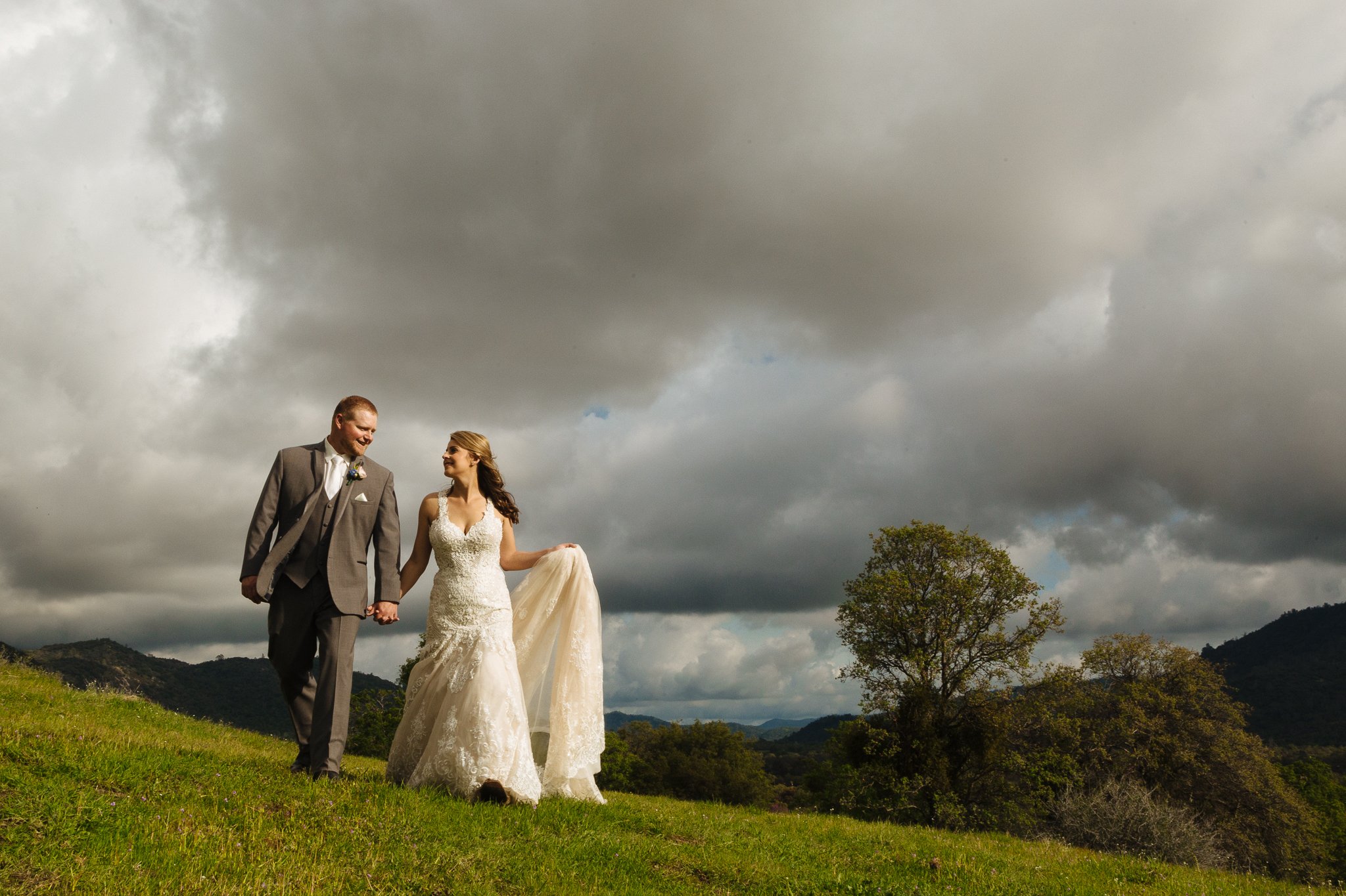 A newlywed couple holding hands and walking on a grassy hill under dark, cloudy skies with mountains and trees in the background.