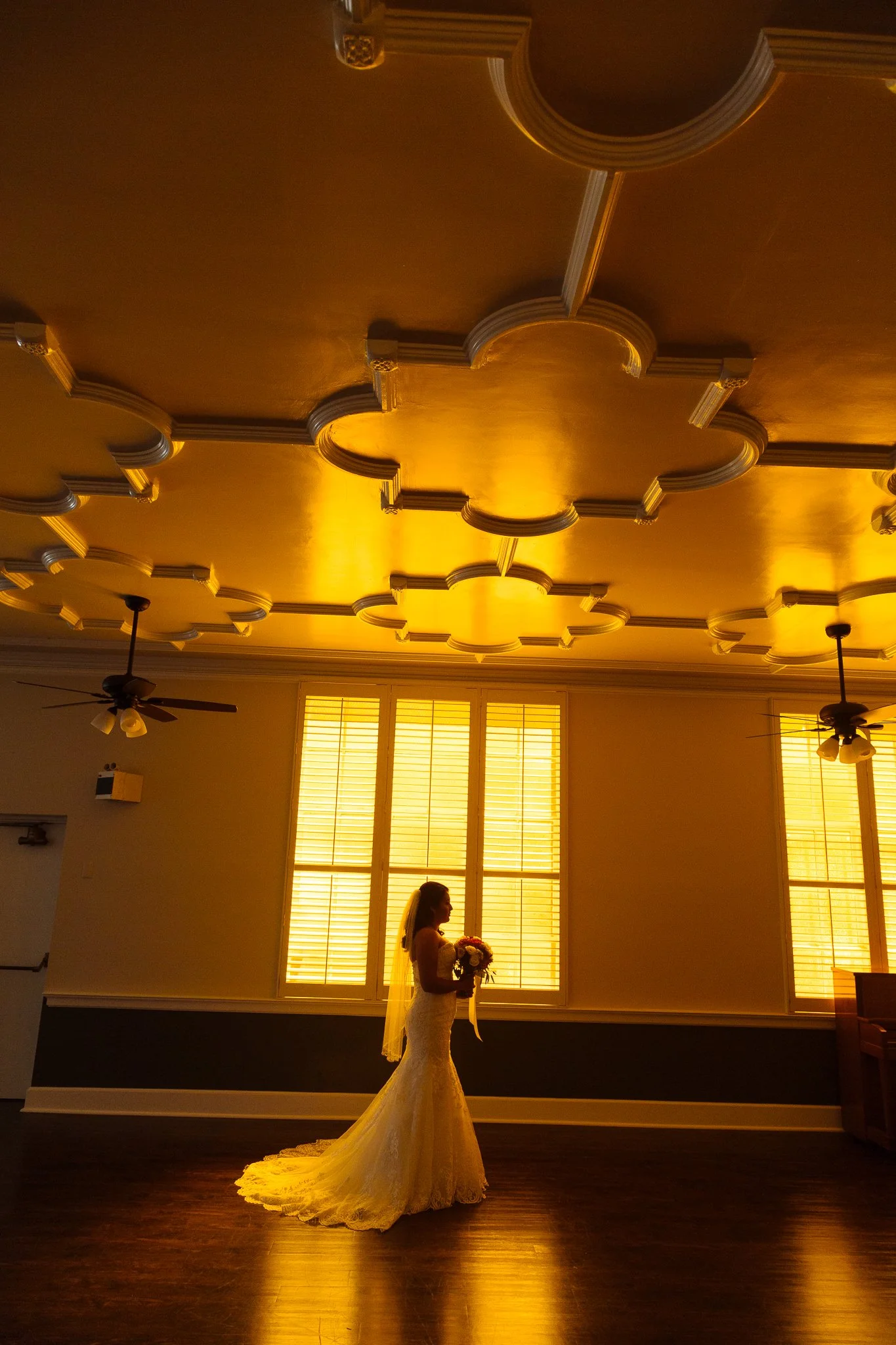 A bride in a wedding gown holding a bouquet, standing in a warmly lit room with large windows and intricate ceiling details.