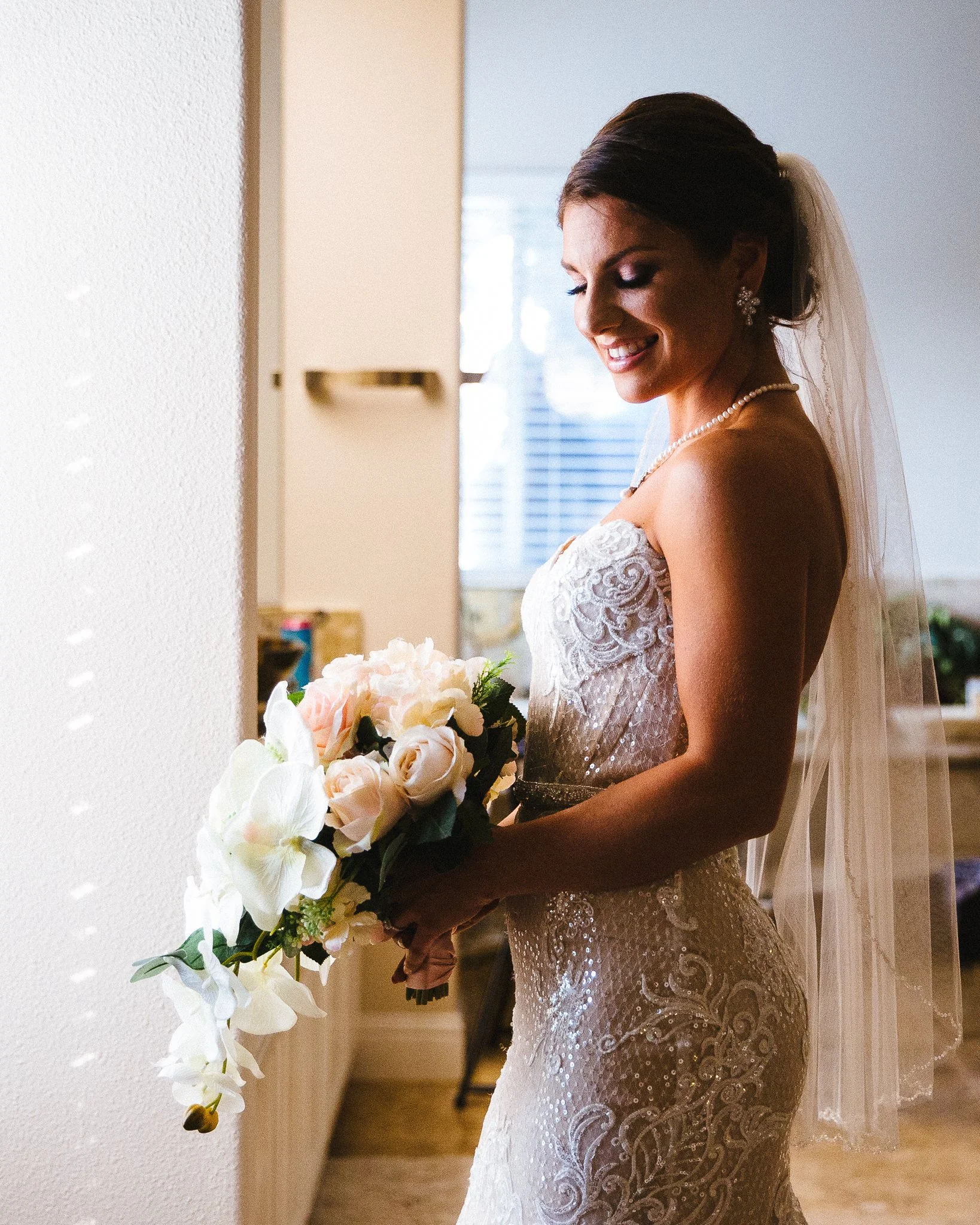 A bride in a wedding dress holding a bouquet of white and pink flowers, with a smile, standing indoors.