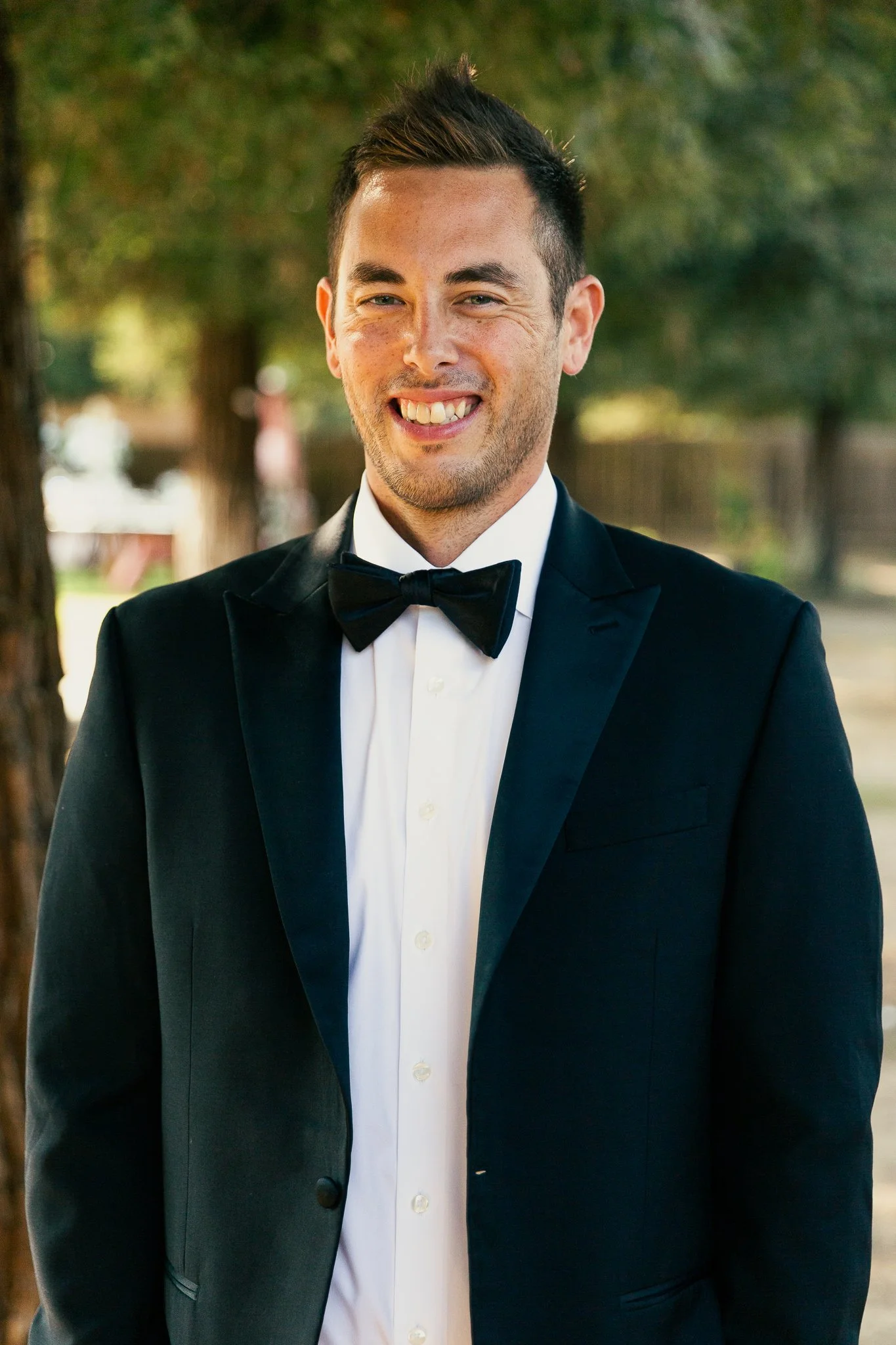 A young man in a tuxedo with a black bow tie, white shirt, and black suit, smiling outdoors with trees in the background.