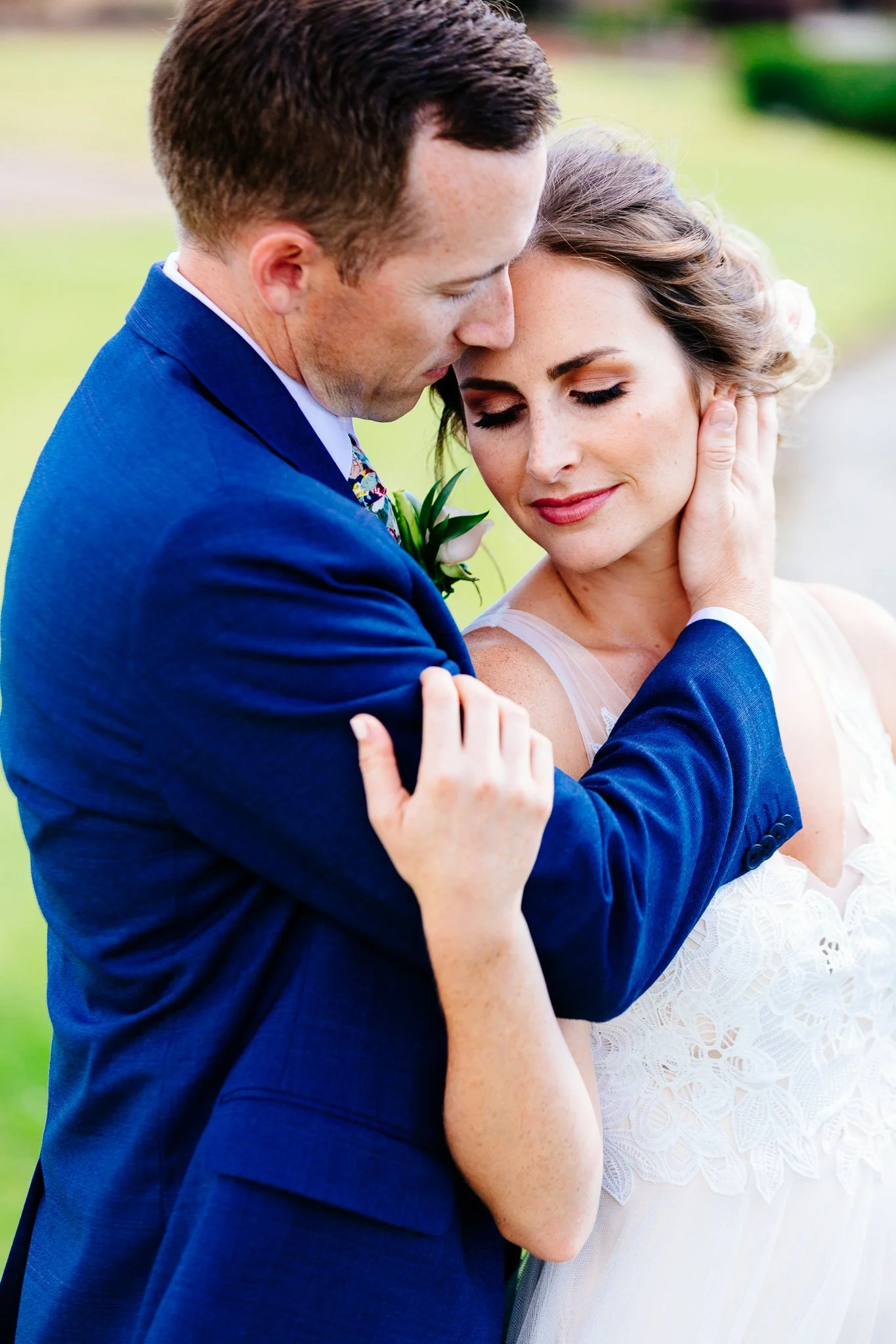 A man and woman embracing outdoors, the man in a blue suit and the woman in a white dress, with their eyes closed and faces touching.