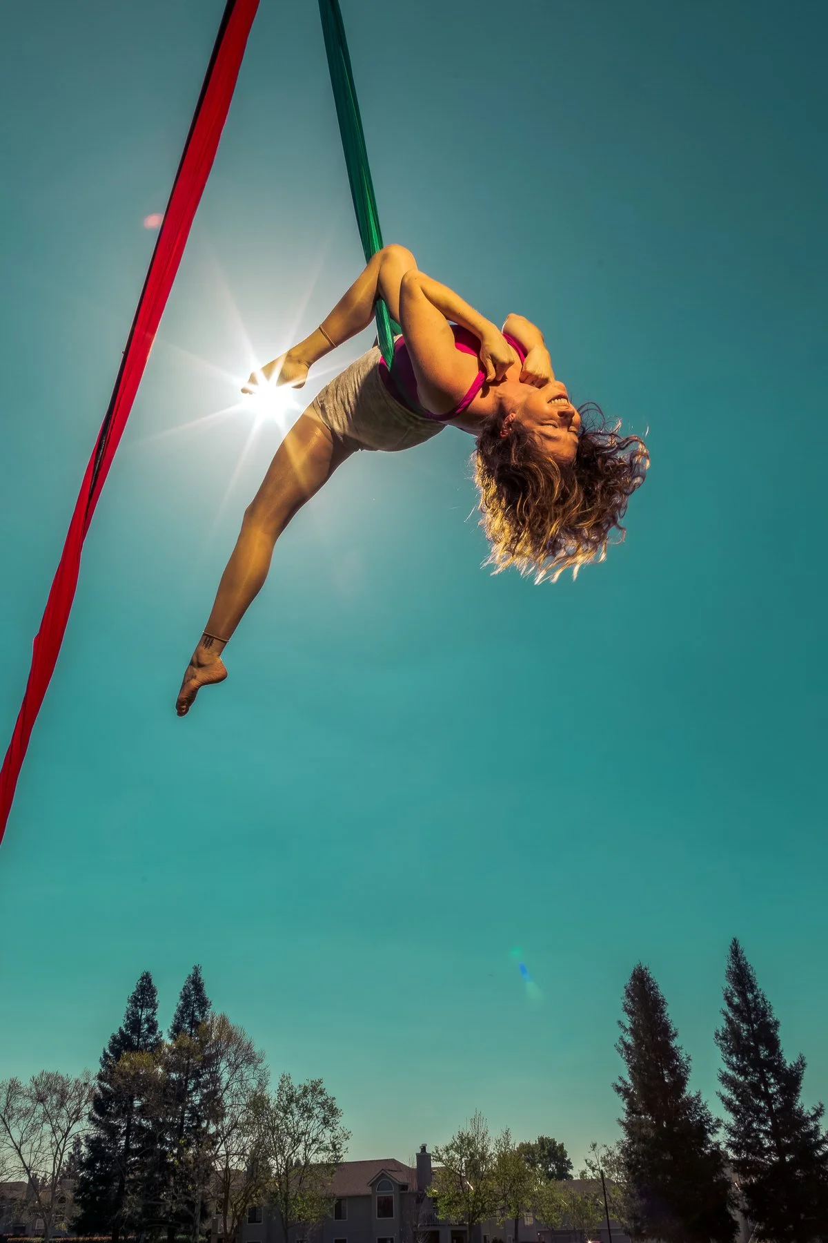 A woman performing an aerial silk pose in the sky, with one hand holding a green silk and the other gripping her shoulder, sunlight shining through her hair, trees and houses visible below, and a clear blue sky.