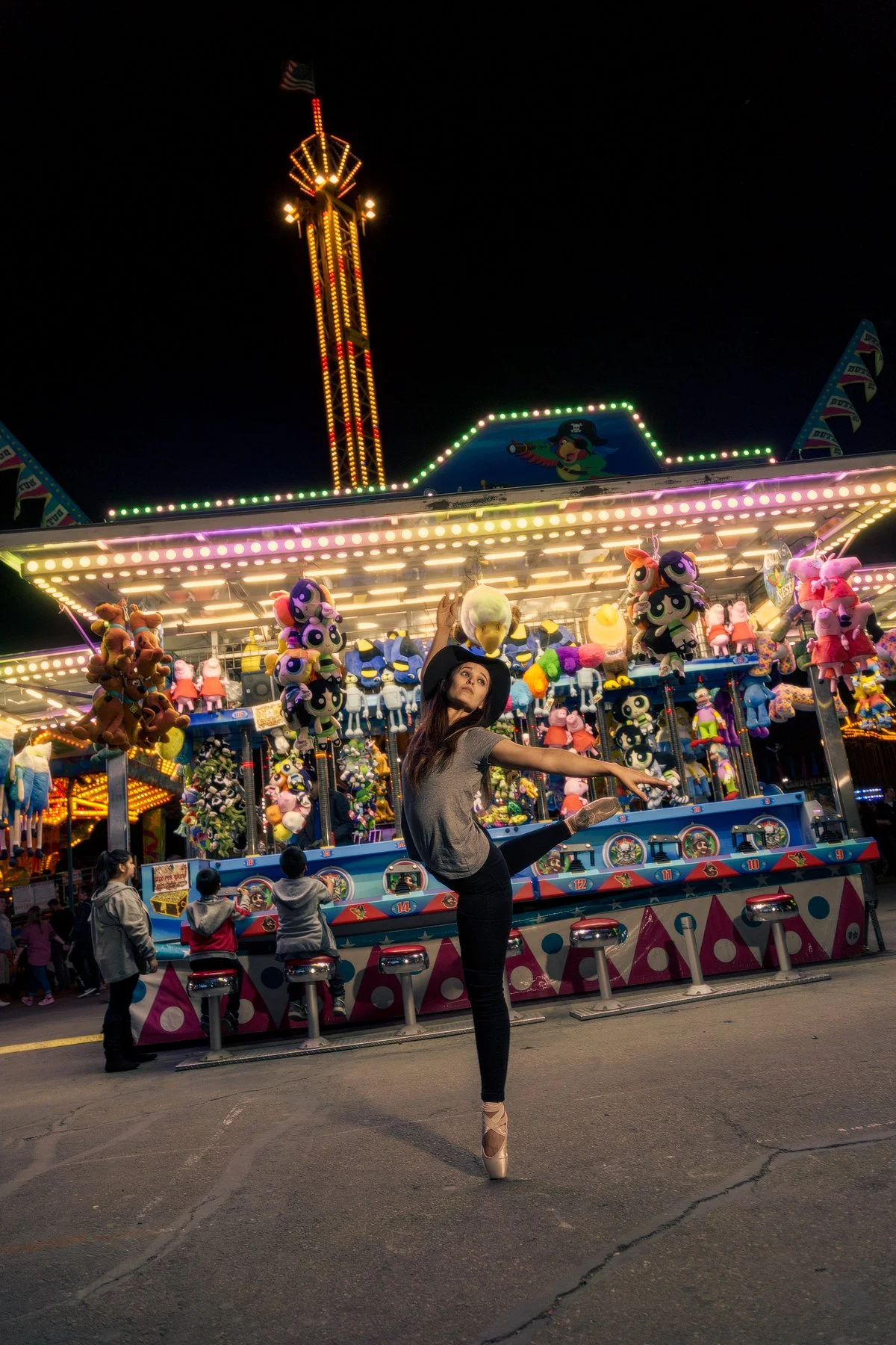 A woman in ballet attire and a black hat dancing in front of a brightly lit amusement park ride at night. The ride is decorated with plush toys and colorful lights, with a few children and other people nearby.