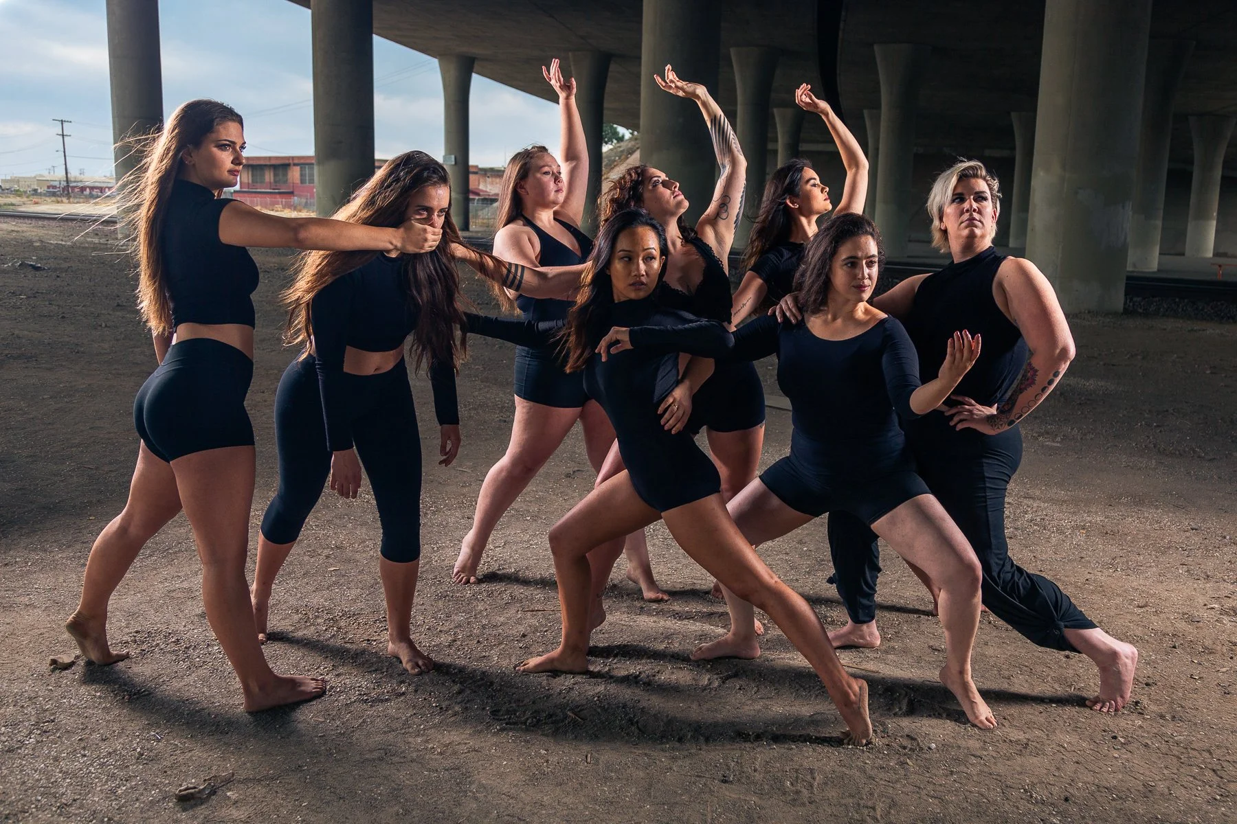 A group of nine women dressed in black performing a dance under a bridge.