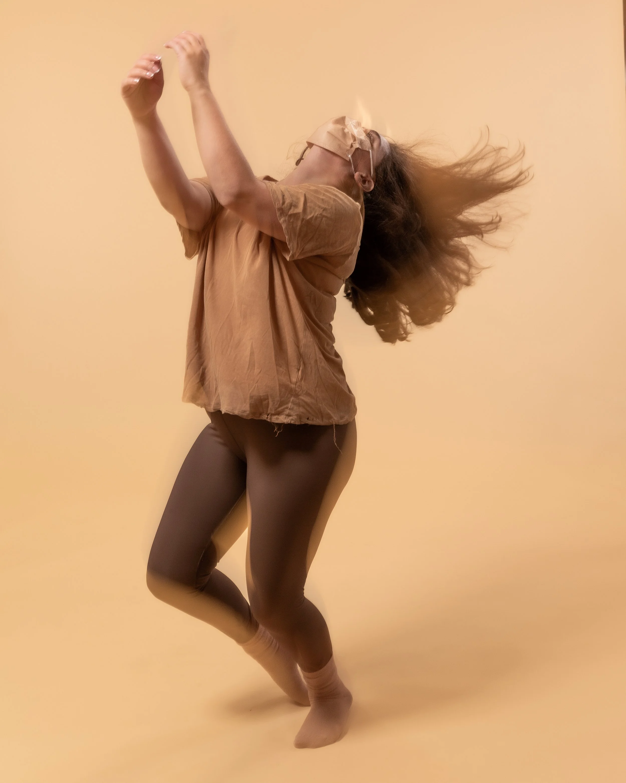 A woman with curly hair dancing against a beige background, wearing glasses, a brown top, and black leggings.