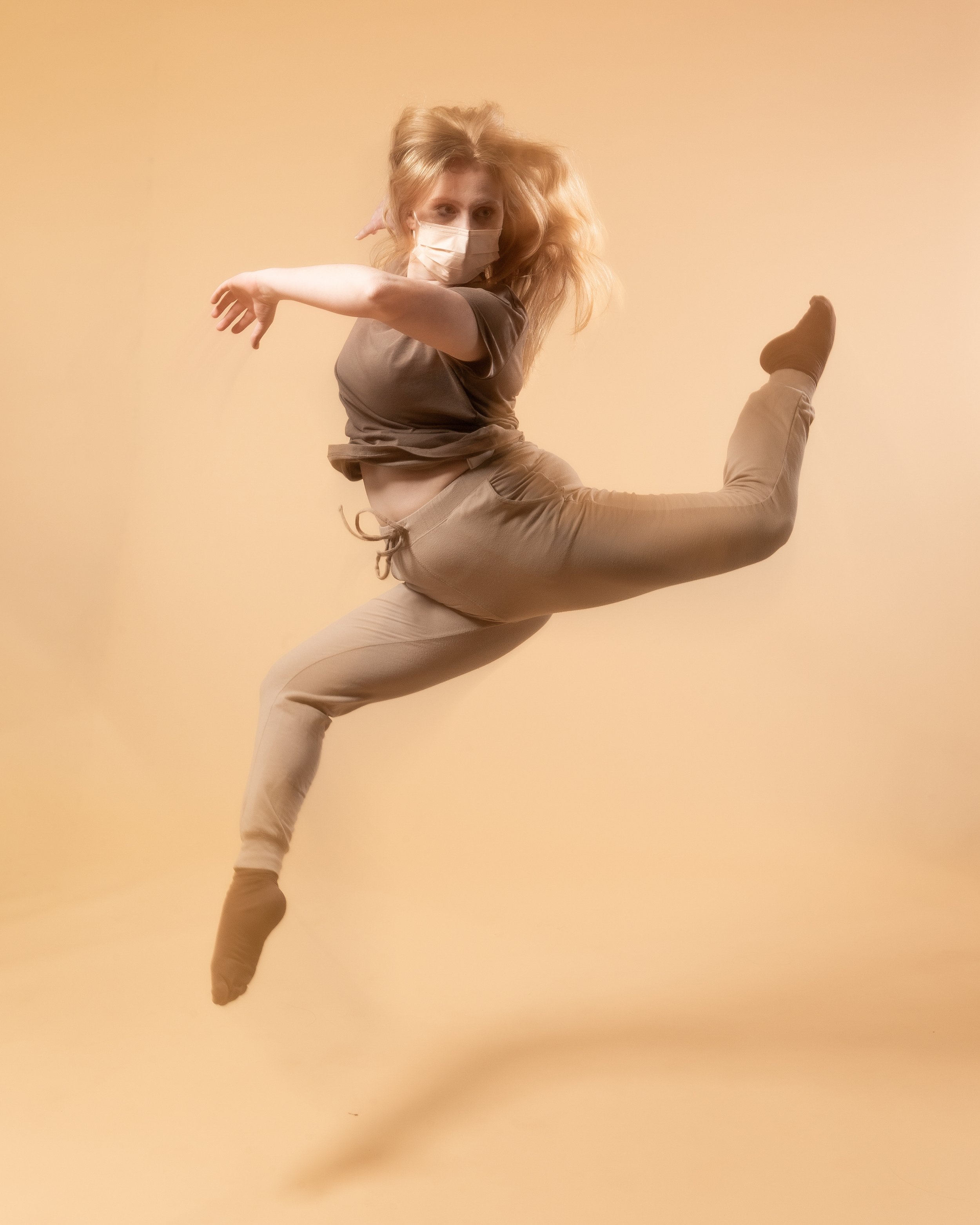 A woman wearing a face mask jumping in mid-air with one leg extended forward and the other bent back, against a plain beige background.