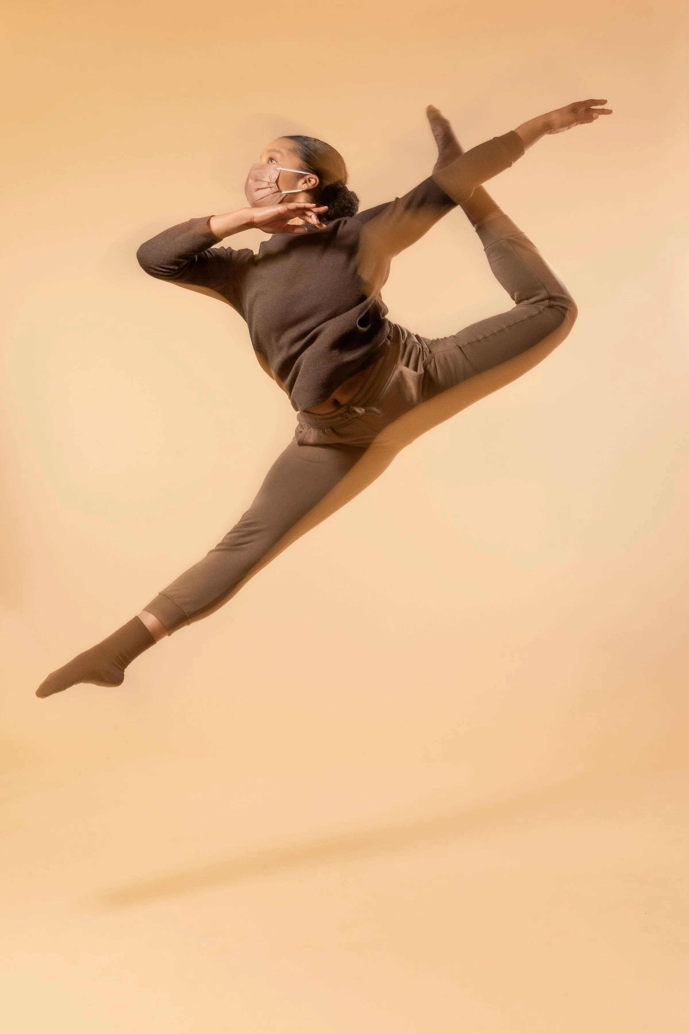 A woman dressed in brown poses mid-air against a beige background, performing a dance or acrobatic move with one leg extended and arms positioned creatively, wearing a face mask.