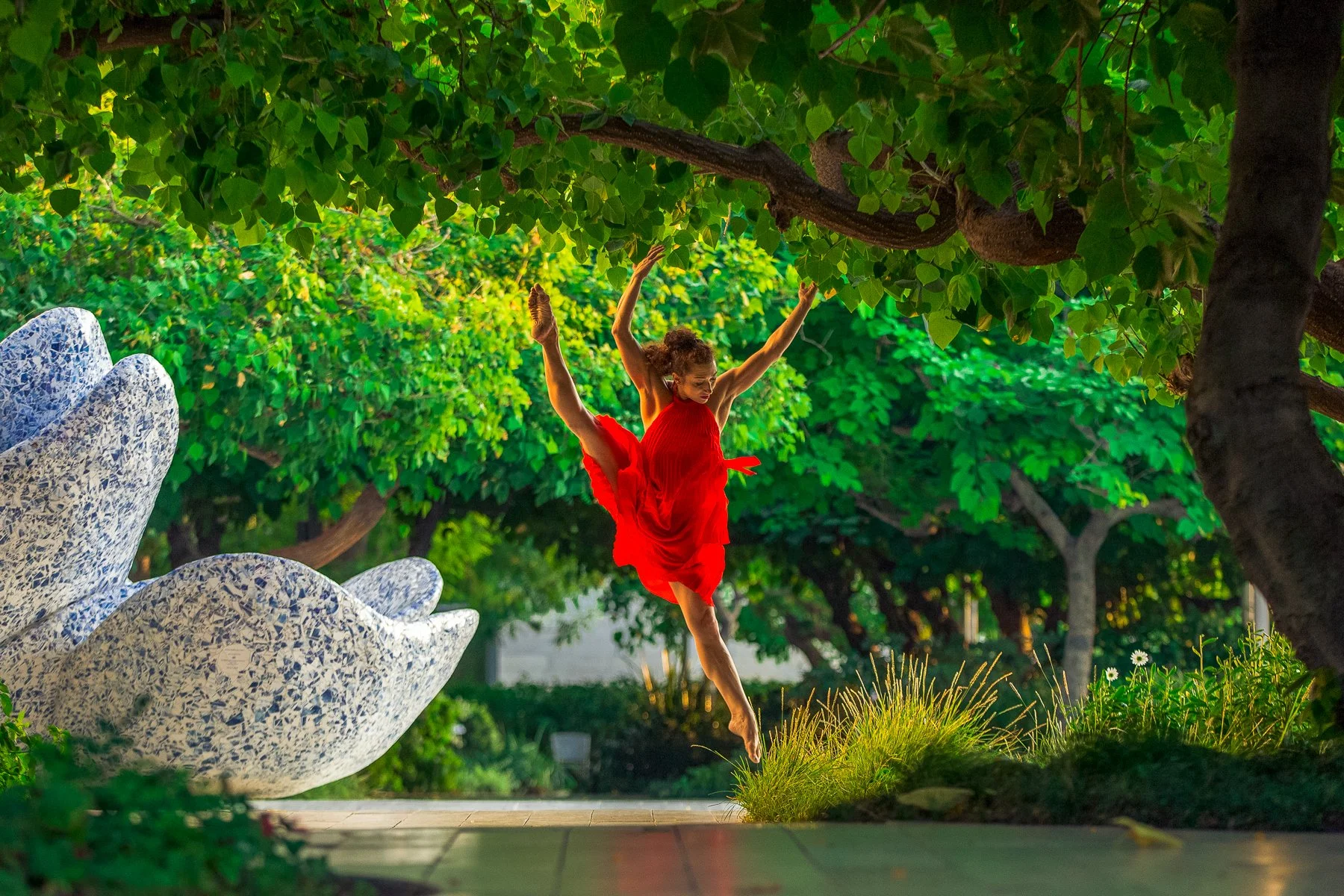 A woman in a red dress dancing or jumping outdoors on a green grassy area, surrounded by lush trees and shrubs, with a decorative stone bench nearby.