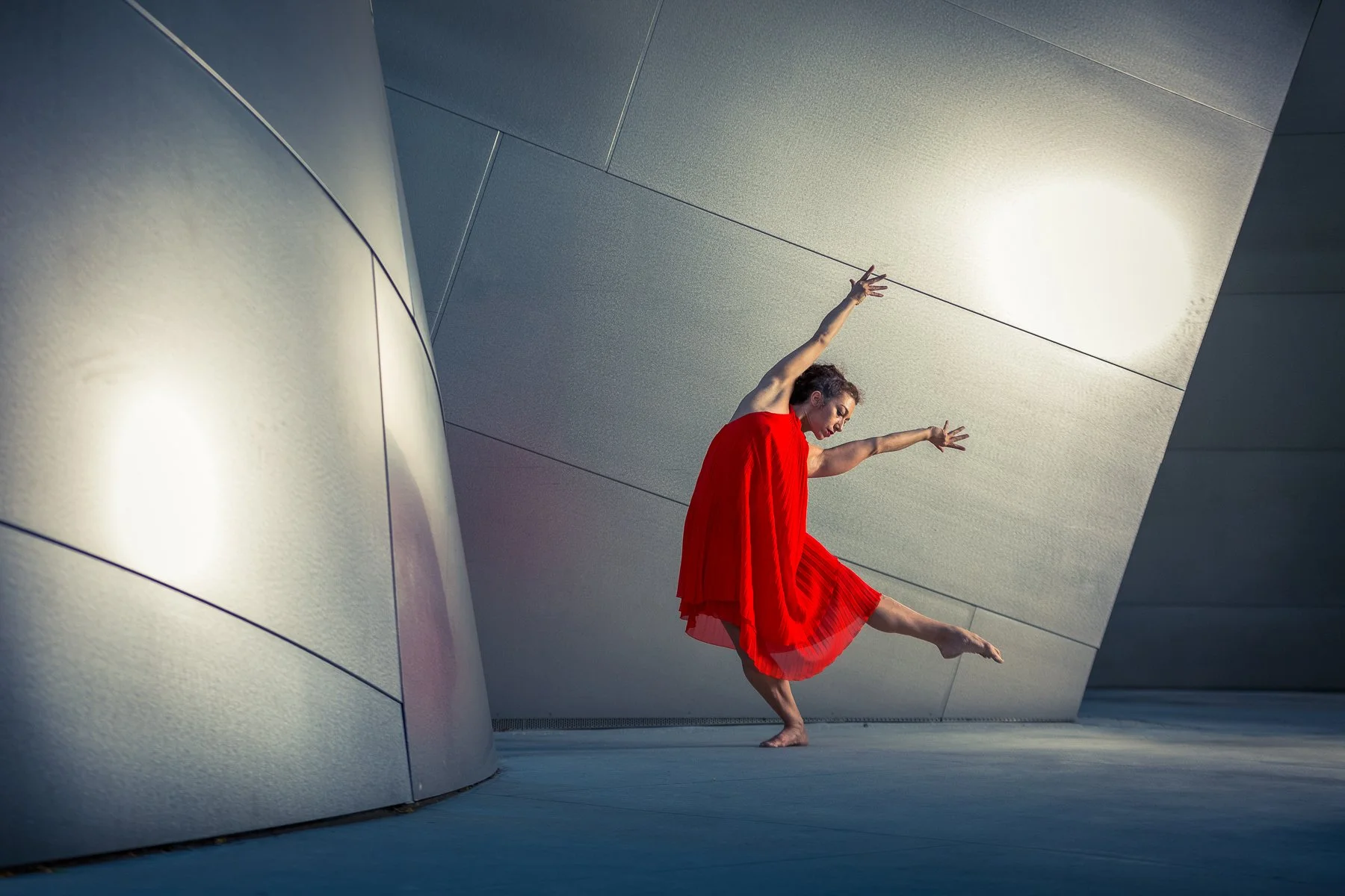 A woman in a red dress dancing in an artistic pose against a modern architectural background with curved and angular metallic surfaces and lights.