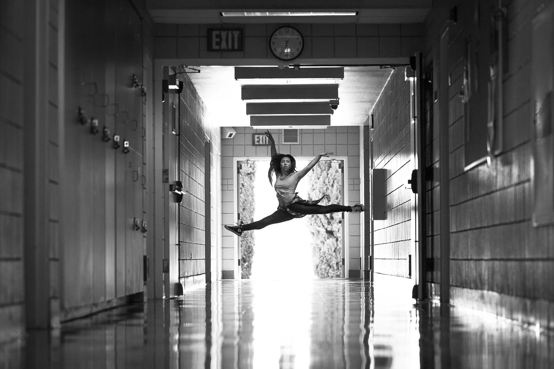 A young woman in athletic clothes performing a high leap in the hallway of a school or gym, with lockers on one side and doors on the other, in black and white.