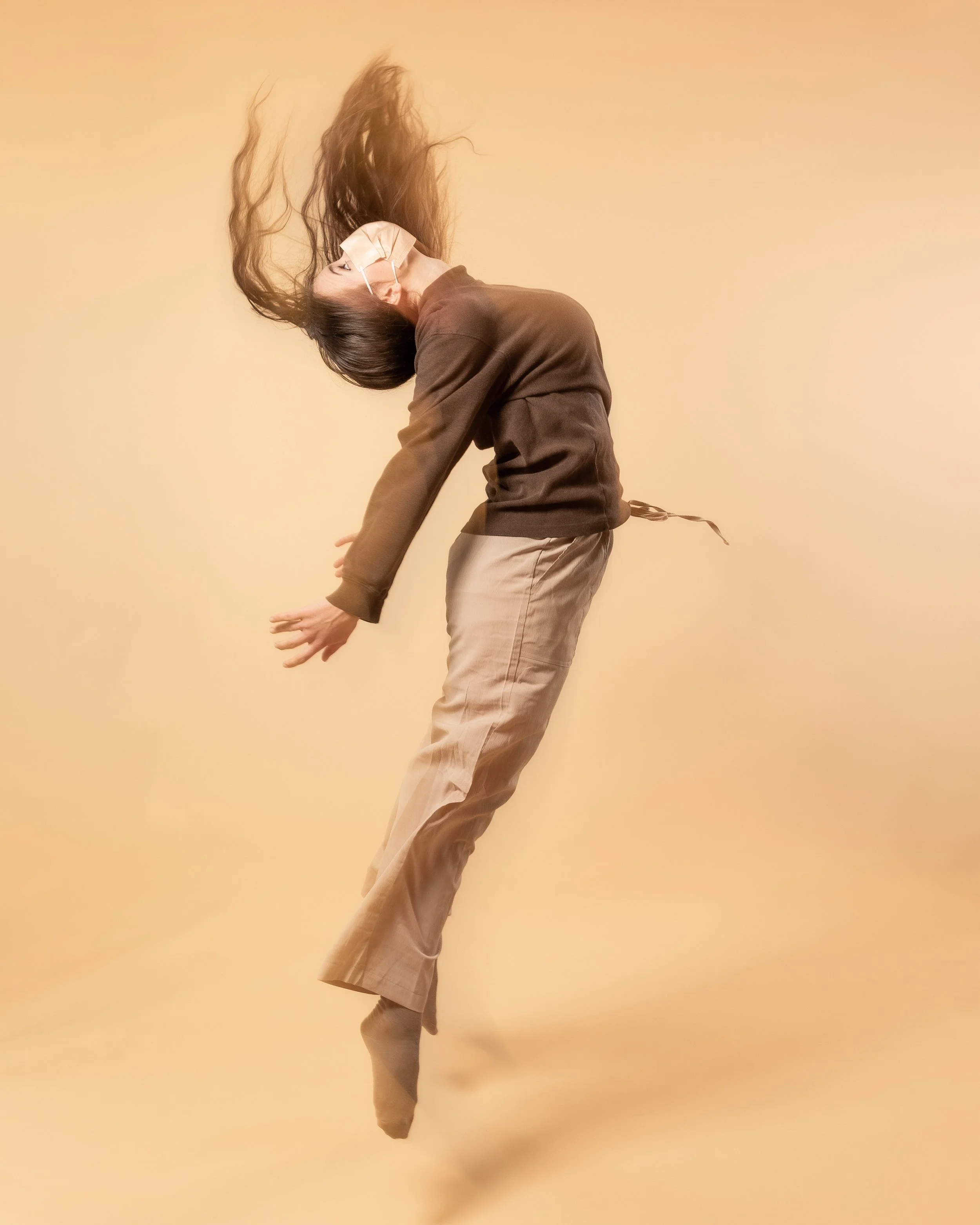 Person floating in mid-air with hair and clothes flowing, against a plain beige background.