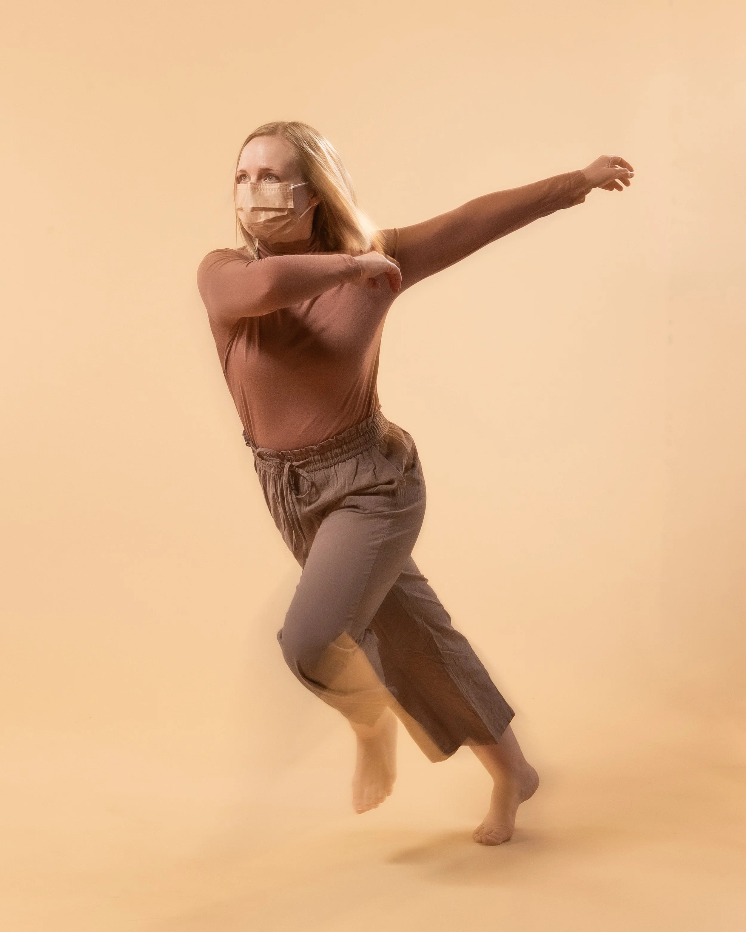 Woman with blonde hair wearing face mask, beige top, and drawstring pants, seen mid-dance move against a solid light beige background.