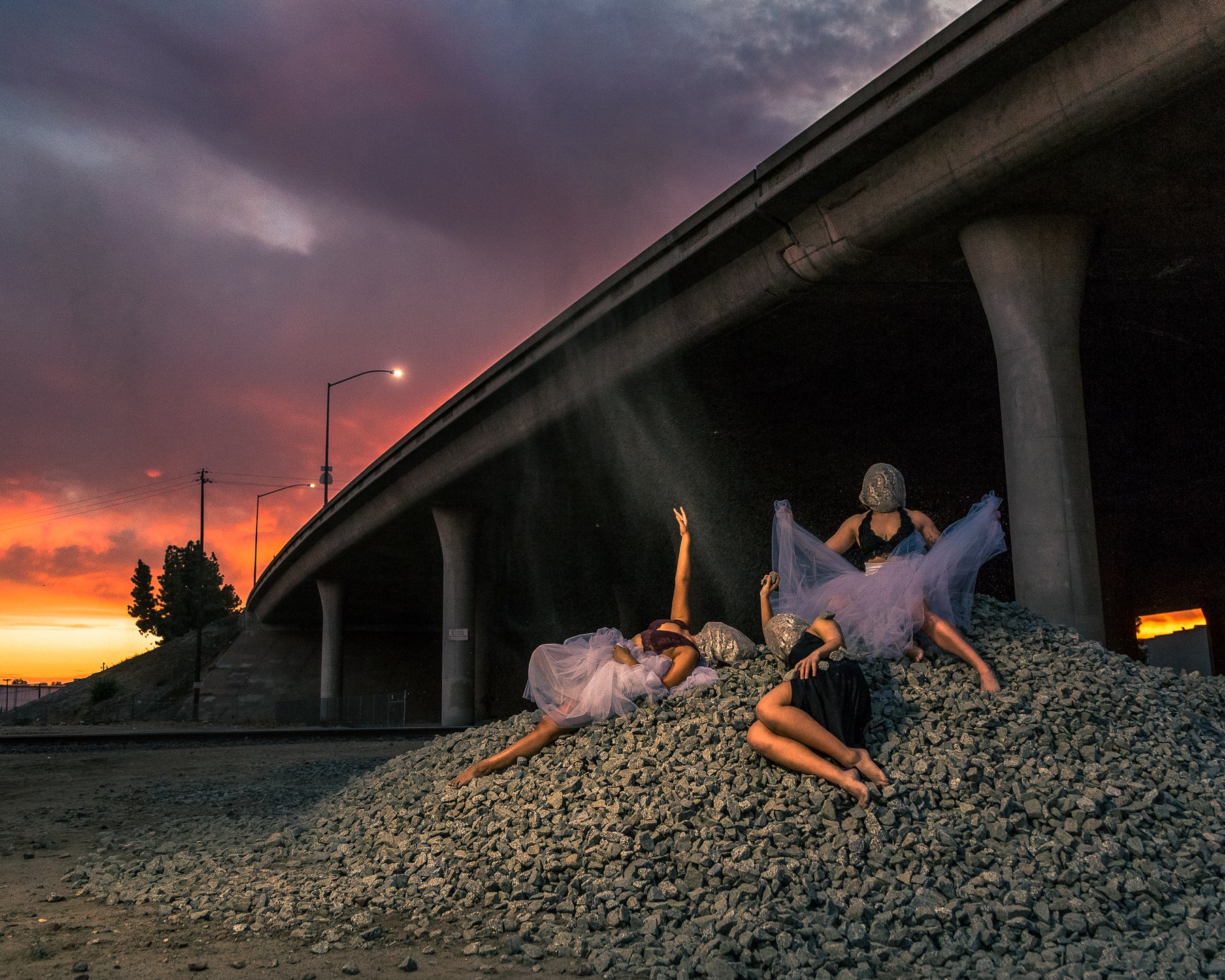 Four women in ballet costumes and masks posing on a pile of rocks under a bridge during sunset.