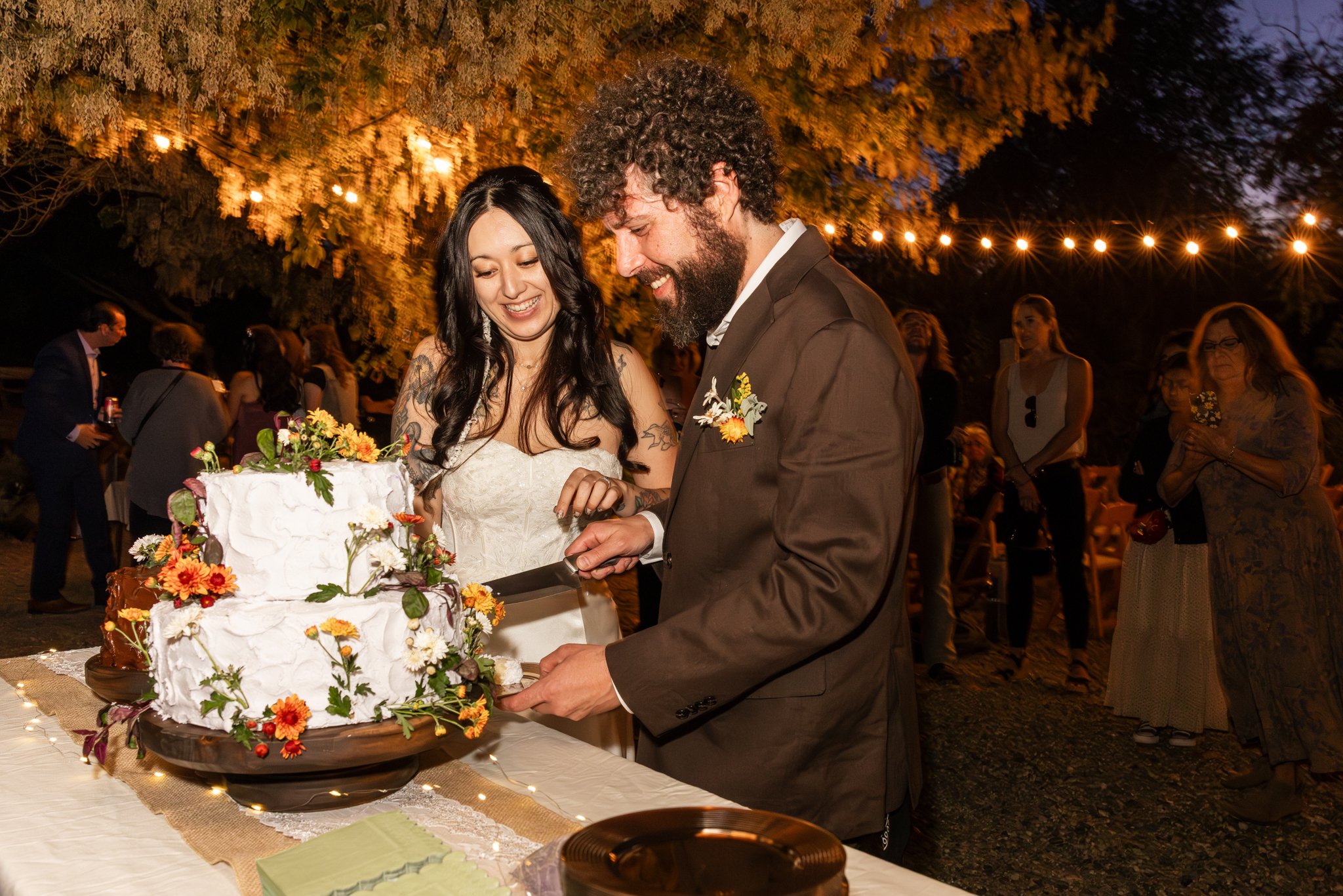 A couple is cutting a wedding cake decorated with orange and yellow flowers at an outdoor evening reception with string lights and guests in the background.
