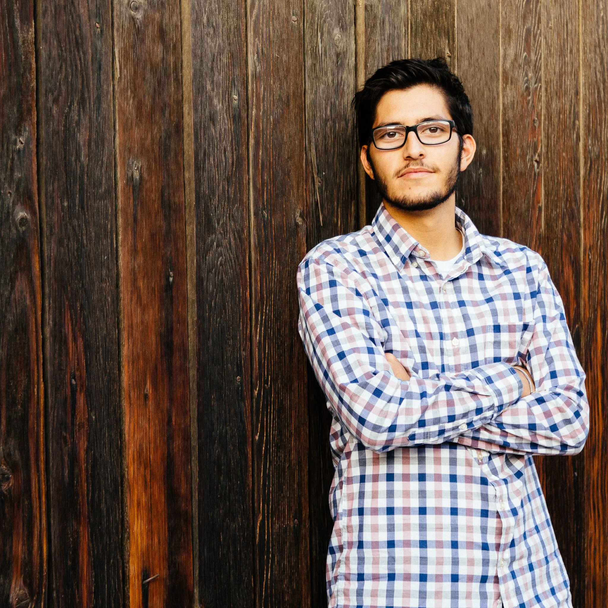 A young man with dark hair, glasses, and a beard, wearing a plaid shirt, stands with arms crossed against a wooden wall.