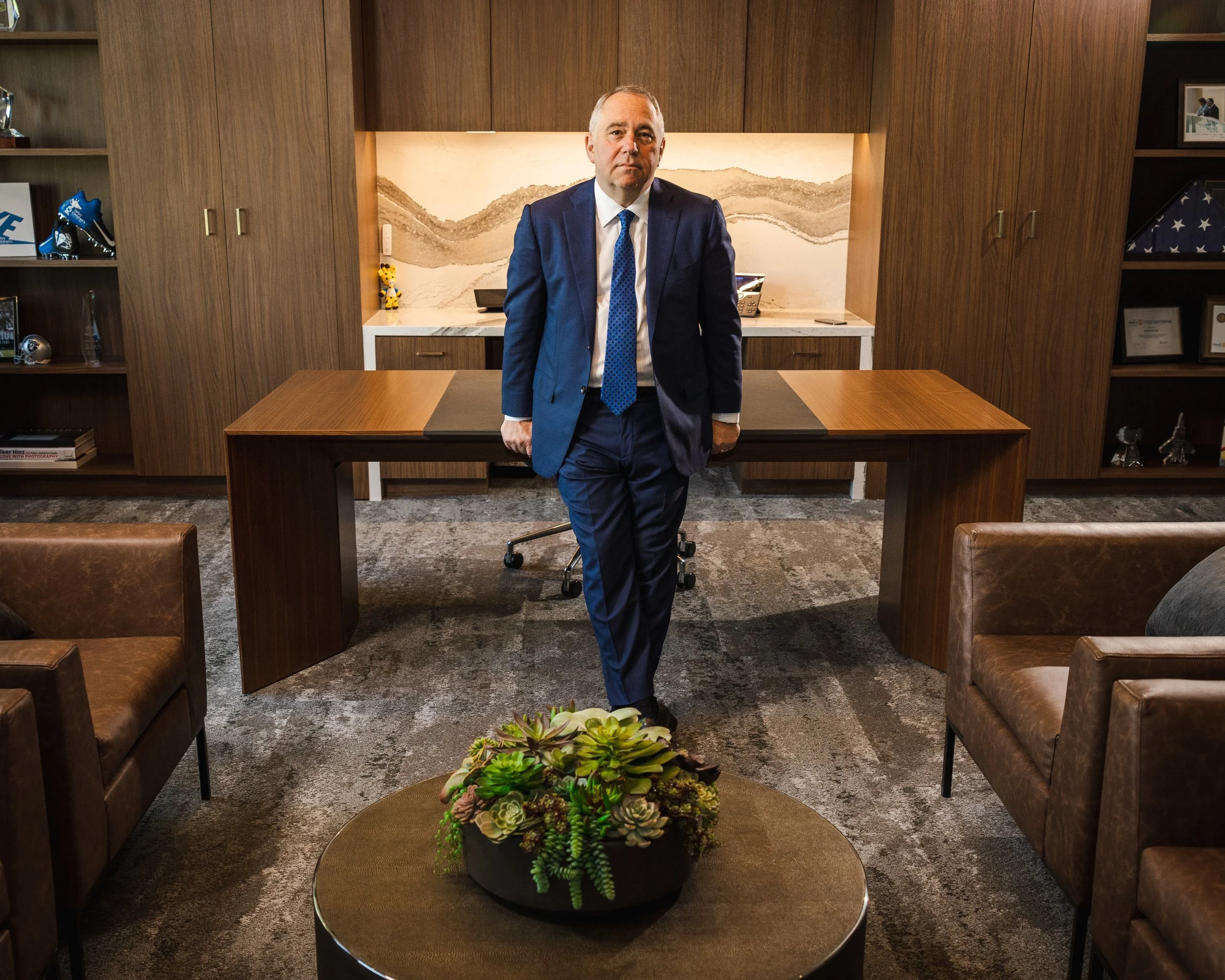 A man in a blue suit standing in a modern office with a wooden desk and bookshelves, and seating area with a round table and couch.