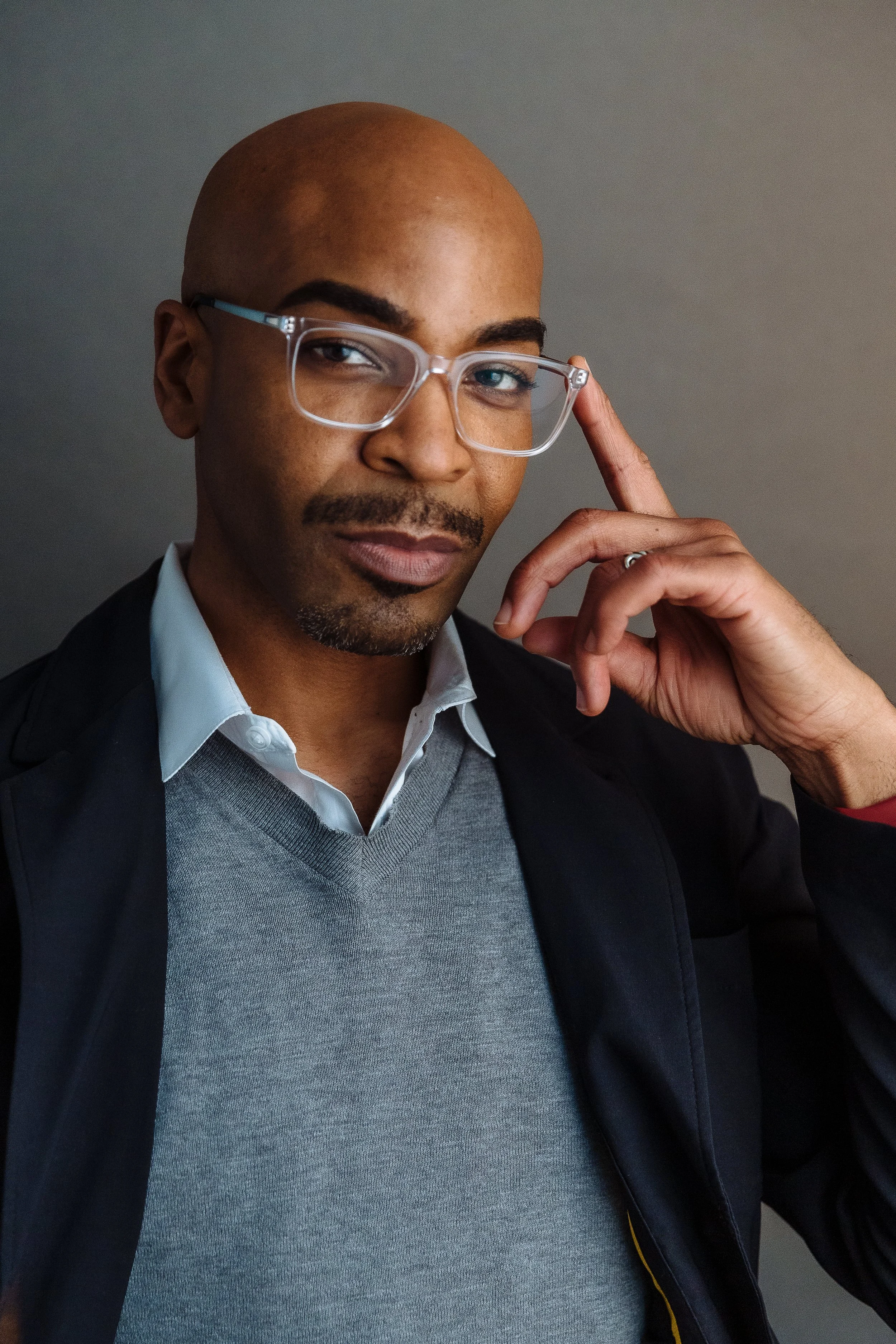 A confident man with glasses, a mustache, and a goatee, dressed in a white shirt, grey sweater, and dark jacket, adjusts his glasses and looks at the camera.