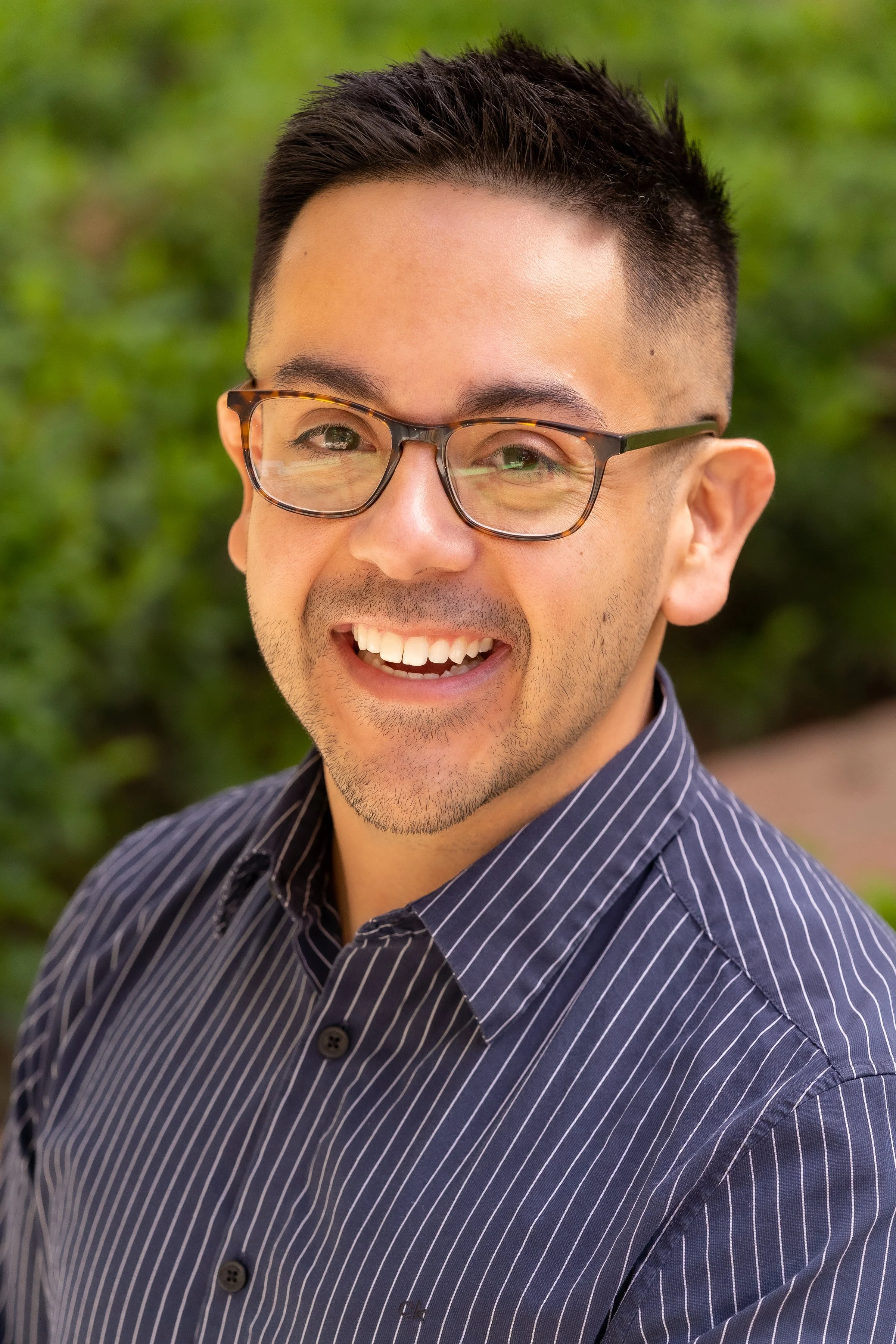 Smiling man with glasses and short dark hair, wearing a striped shirt, outdoors with green foliage in the background.